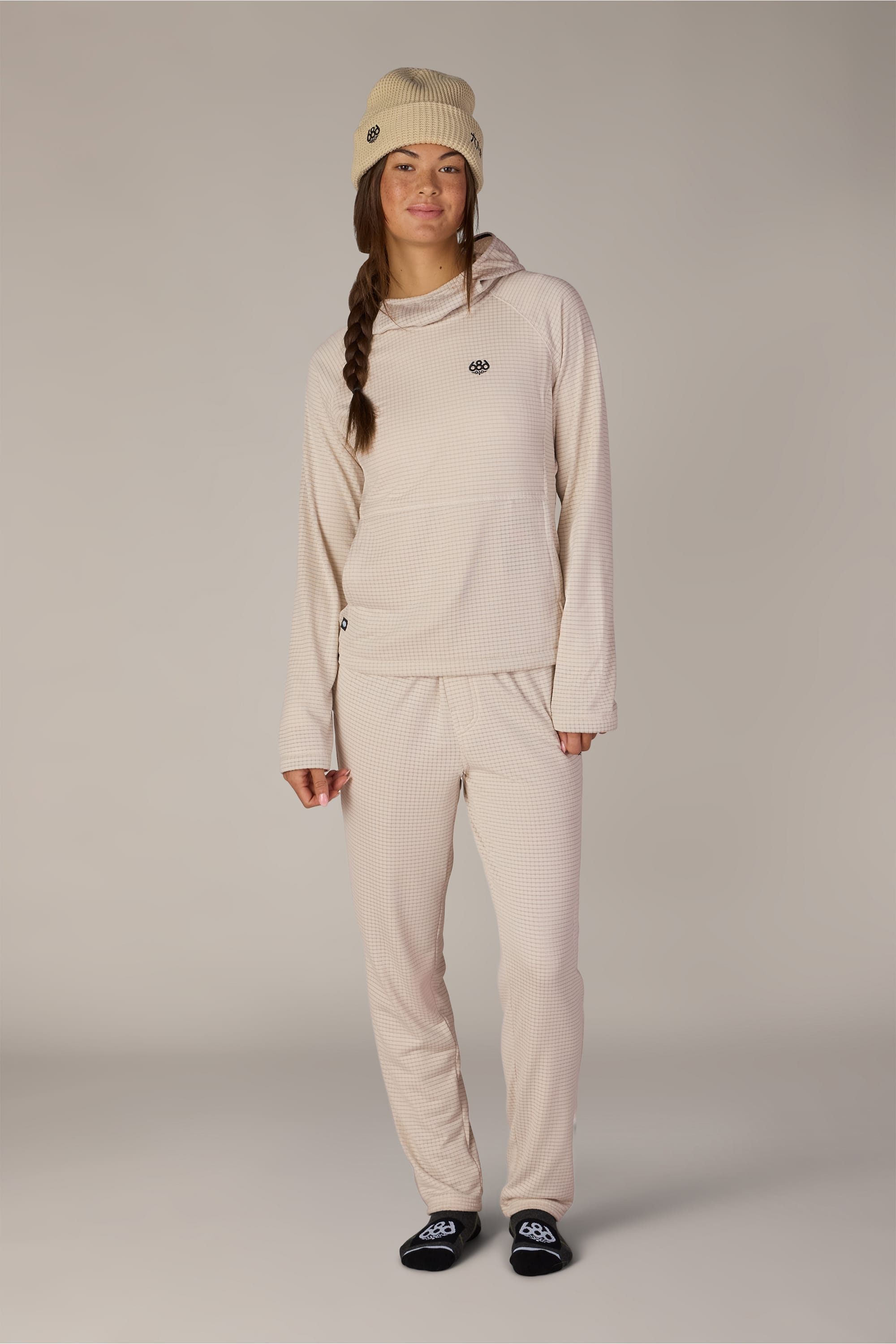 A person stands and smiles against a plain light background, effortlessly showcasing the comfort and style of the 686 Women’s Mission Grid™ Hoody by 686, paired with matching beige pants and beanie, as well as black socks.