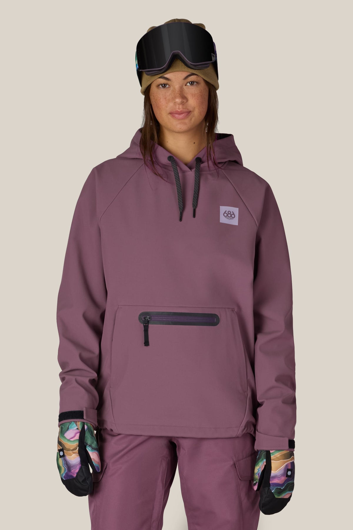 A person wearing the 686 Women’s Waterproof Hoody in mauve stands facing forward against a plain beige background, paired with matching pants, colorful gloves, a beanie, and ski goggles on their head.