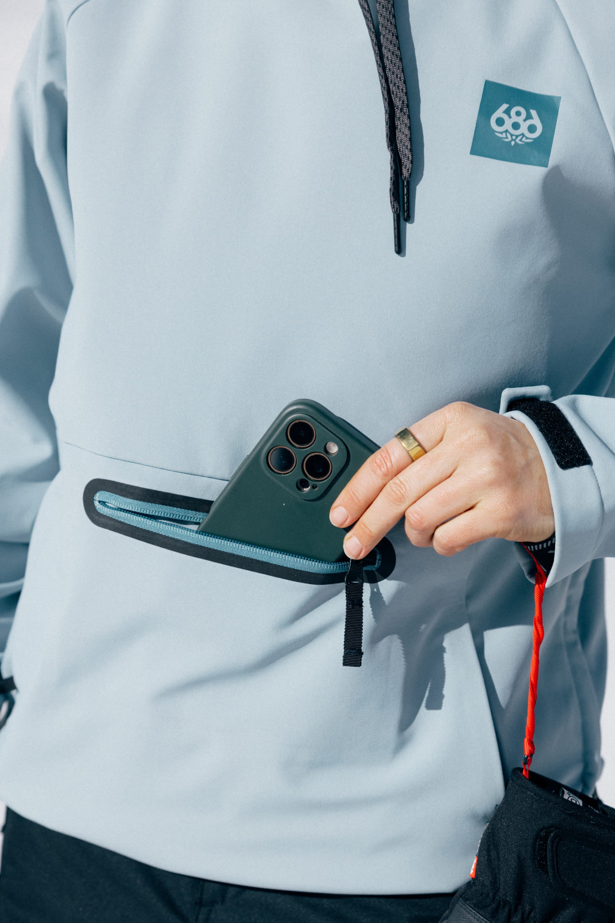 A person wearing the 686 Women’s Waterproof Hoody in light blue places a green smartphone into a zippered pocket, with only their hand and part of the hoody visible.