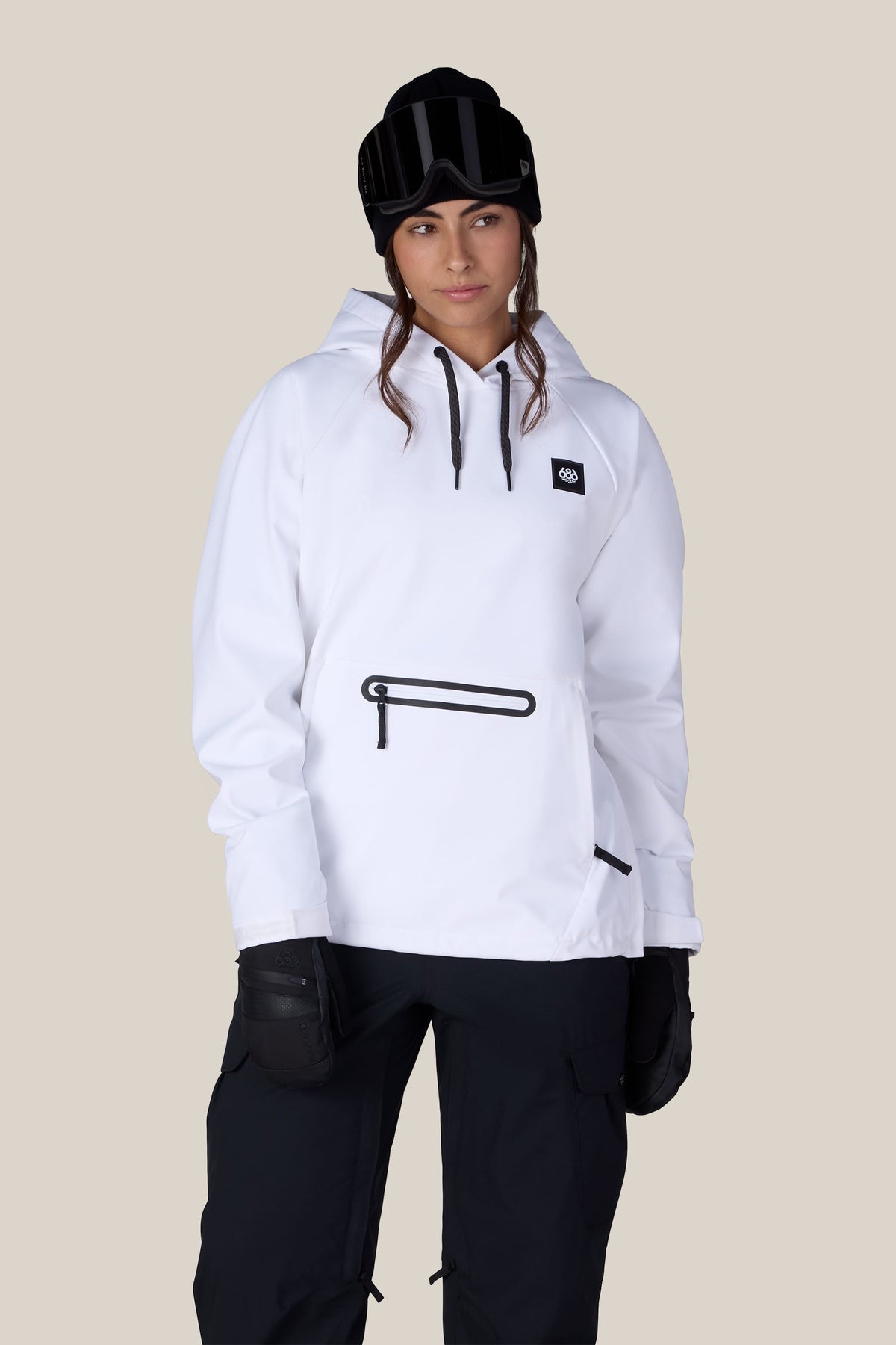 A person wearing the 686 Women’s Waterproof Hoody by 686, paired with black gloves, black pants, and a black beanie with goggles on top, stands against a plain beige background.