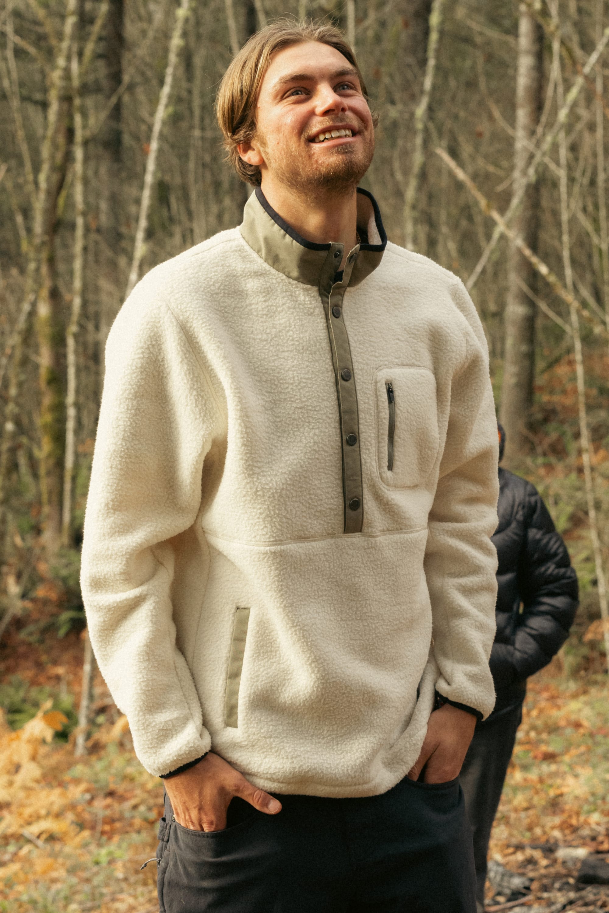 A smiling man wears a cream-colored 686 Mens Tioga Fleece Pullover, standing outdoors in a forest with bare trees and autumn leaves in the background.