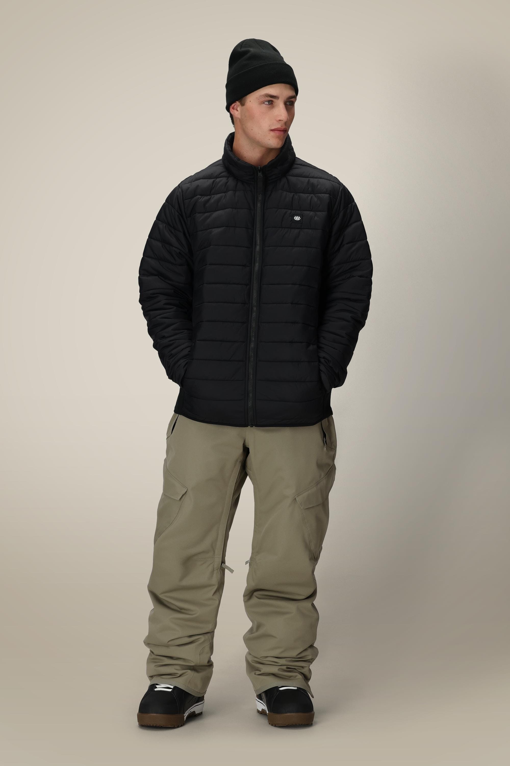 A man wearing a black beanie, the 686 Men’s Thermal Puff Jacket by 686, khaki cargo pants, and black shoes stands with his hands in his pockets, looking to the side against a plain light background.