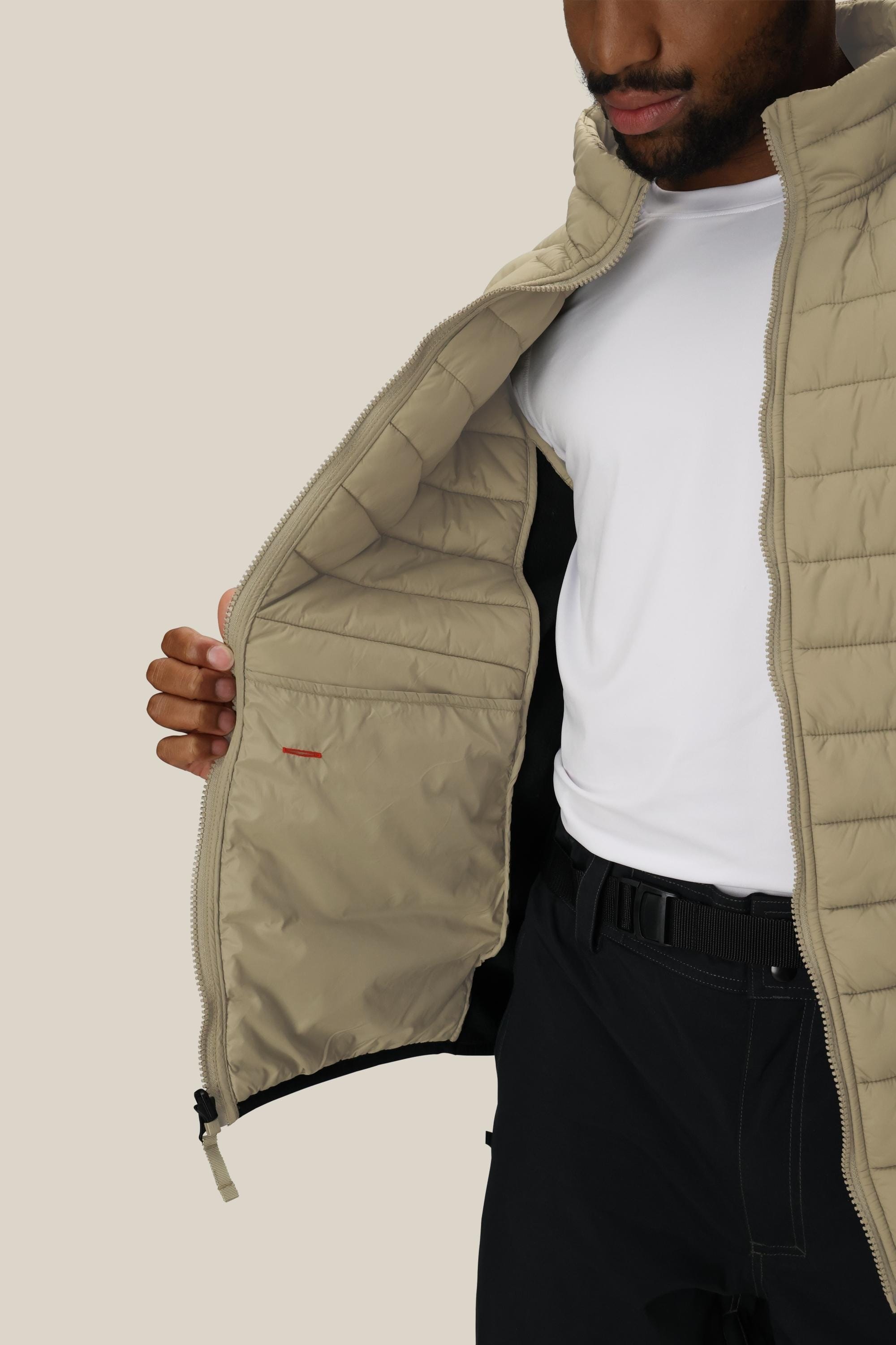 A man wears the 686 Mens Thermal Puff Jacket in beige, holding it open to reveal an inner pocket over his white t-shirt and black pants.