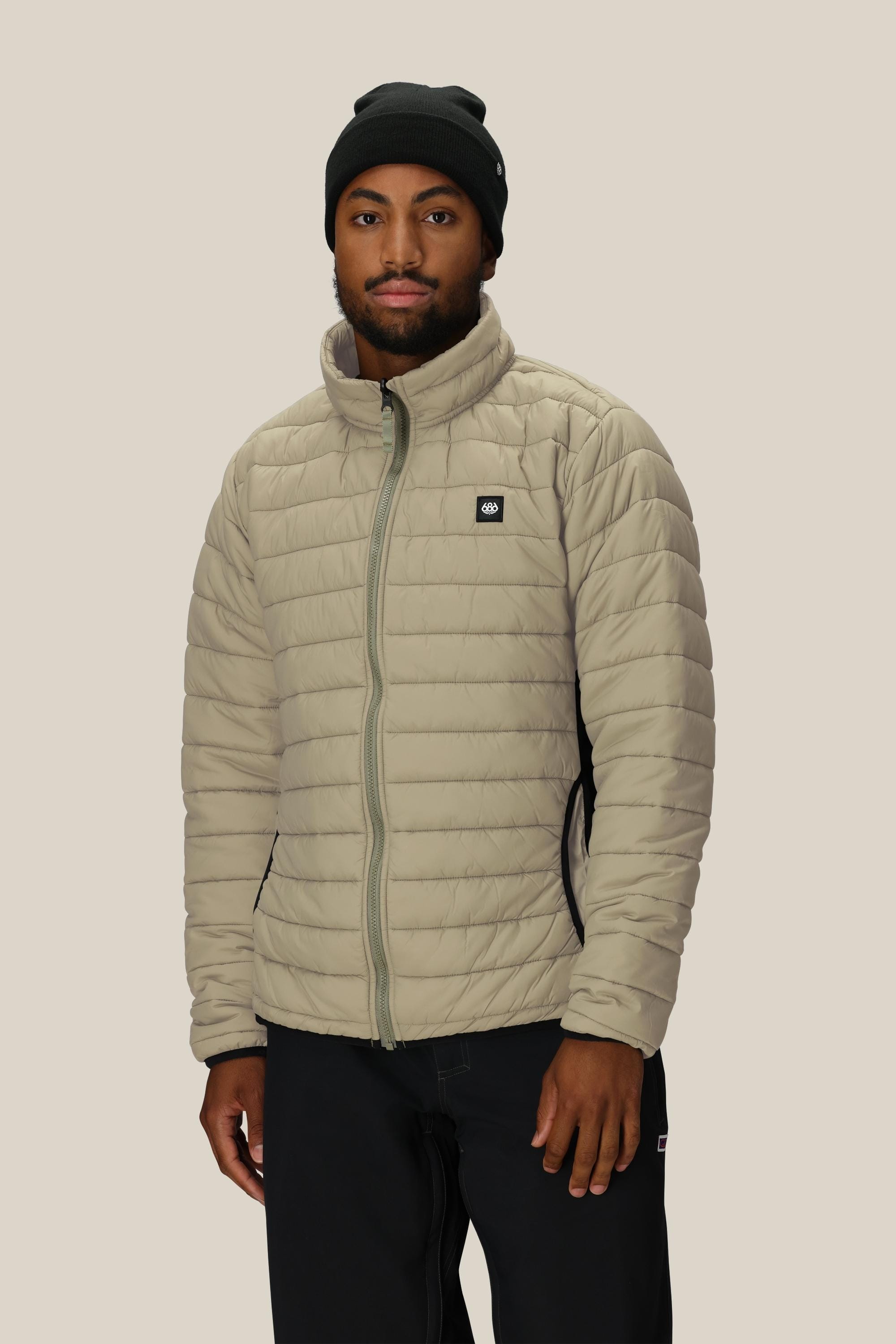 A man wearing the 686 Mens Thermal Puff Jacket in light beige, along with a black beanie and black pants, stands against a plain beige background facing forward with a neutral expression.