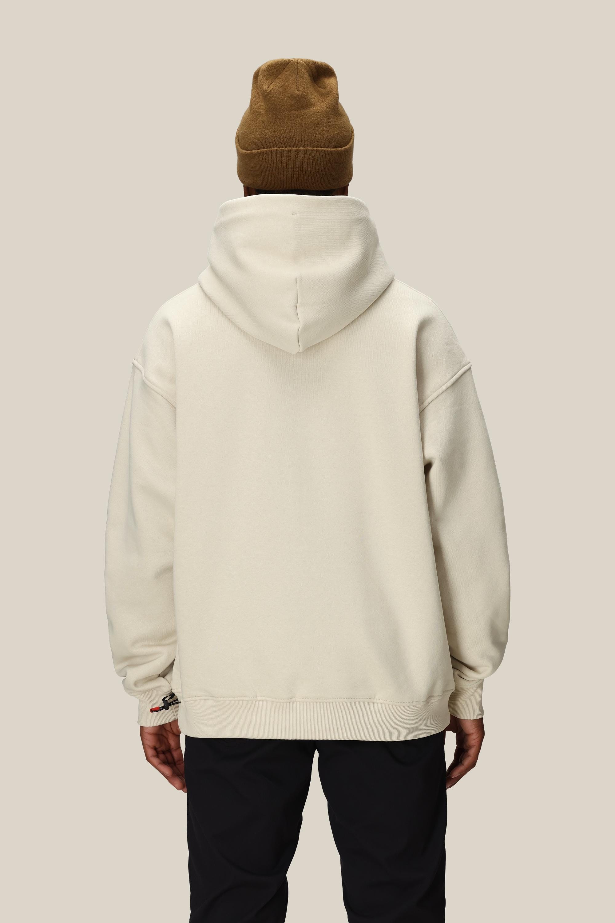 A person wearing a brown beanie, black pants, and the 686 Knockout Logo Premium Heavyweight Pullover Hoody by 686 stands with their back to the camera against a plain beige background.