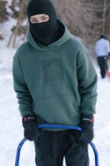 A person in a 686 Knockout Logo Premium Heavyweight Pullover Hoody, black ski mask, and gloves stands outdoors in the snow holding a blue sled handle, with trees and another person visible in the snowy background.