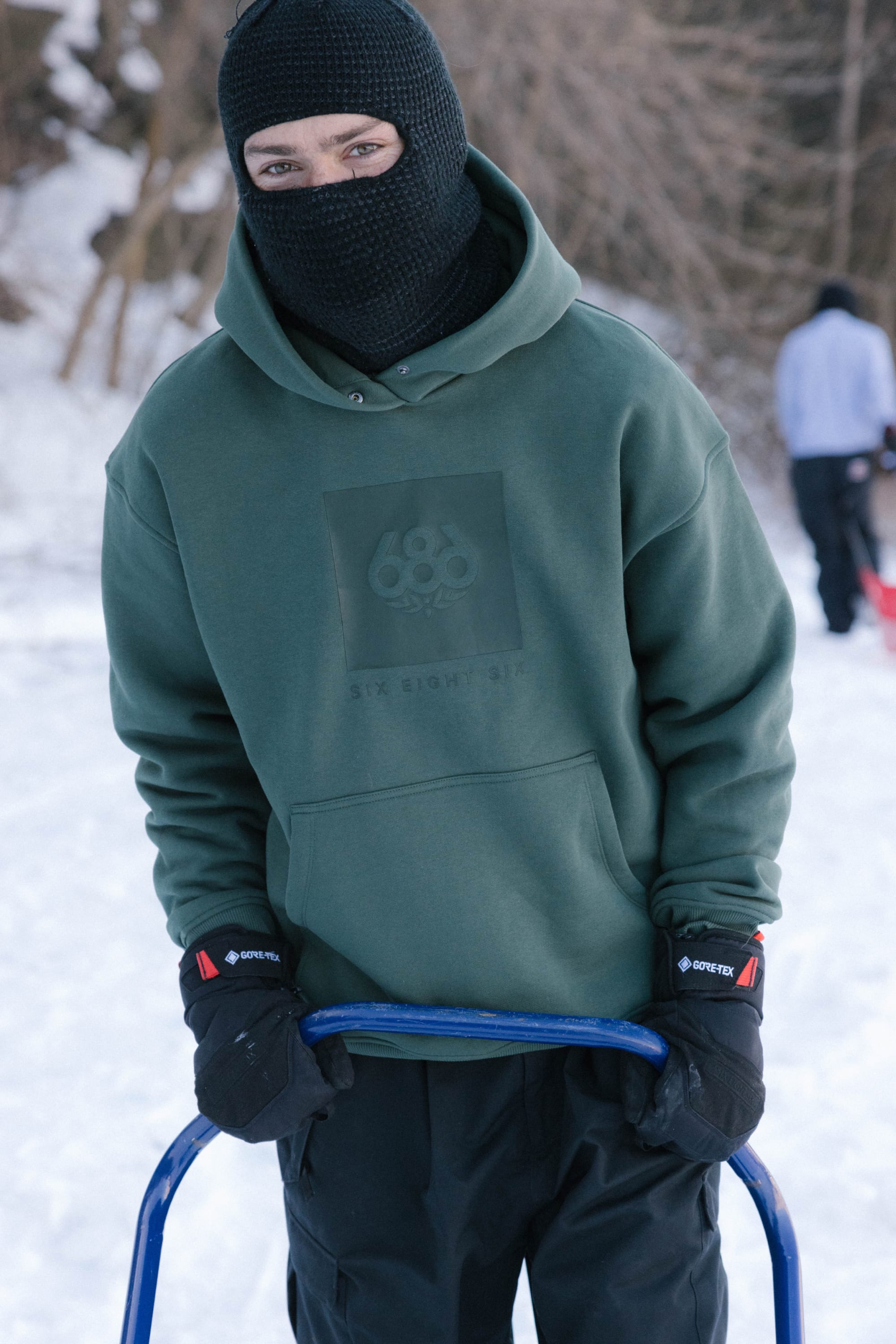 A person in a 686 Knockout Logo Premium Heavyweight Pullover Hoody, black ski mask, and gloves stands outdoors in the snow holding a blue sled handle, with trees and another person visible in the snowy background.