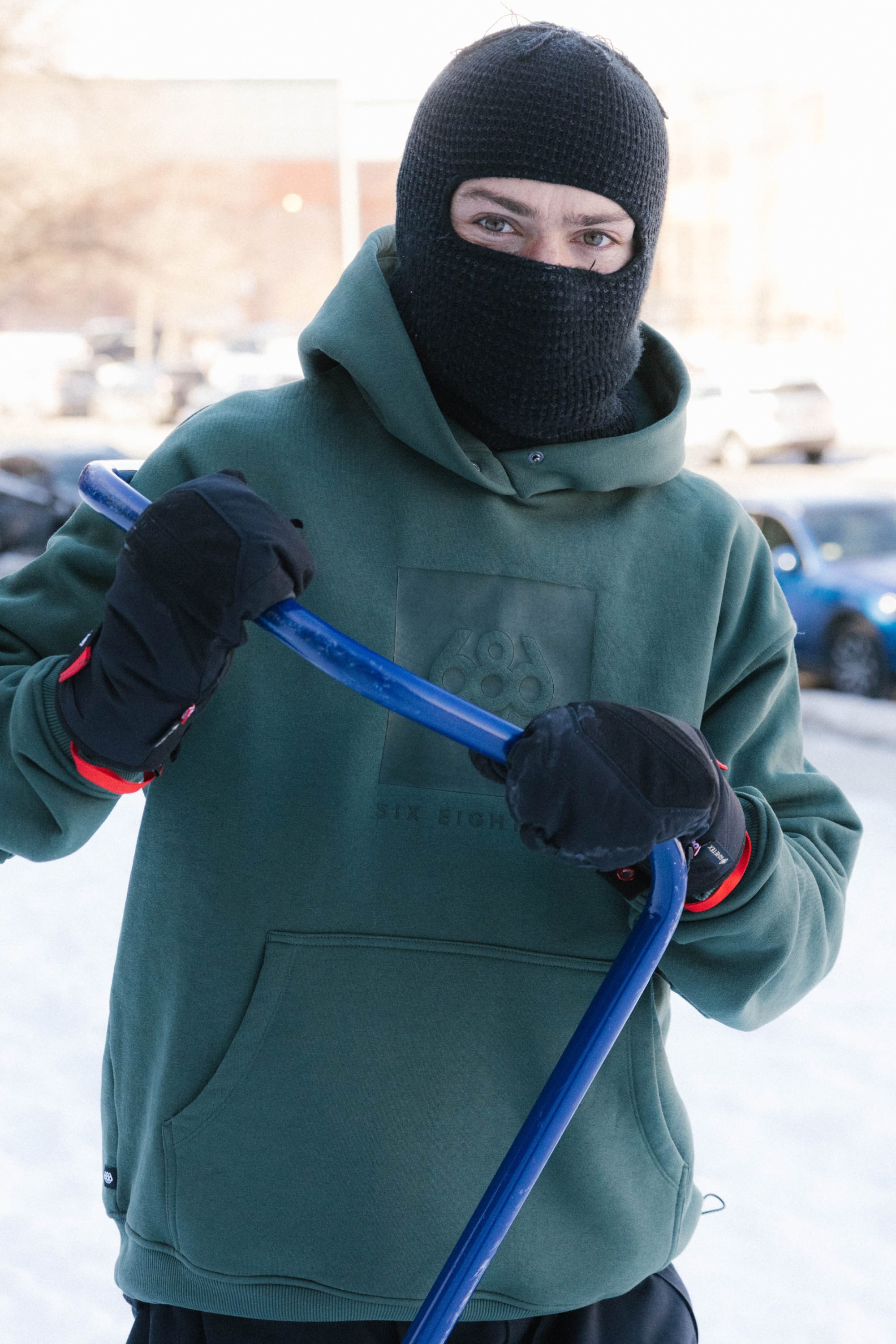 A person wearing a 686 Knockout Logo Premium Heavyweight Pullover Hoody, black ski mask, and black gloves holds a blue snow shovel outdoors on a snowy day, with parked cars visible in the background.