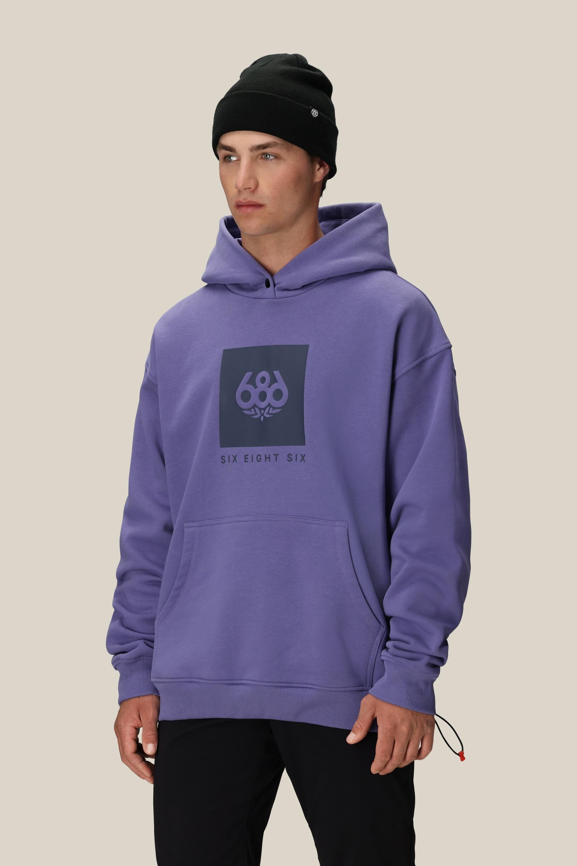 A person wears a black beanie and the 686 Knockout Logo Premium Heavyweight Pullover Hoody—a loose, purple men’s cotton sweatshirt with a front pocket and “686” chest logo—standing against a plain beige background.
