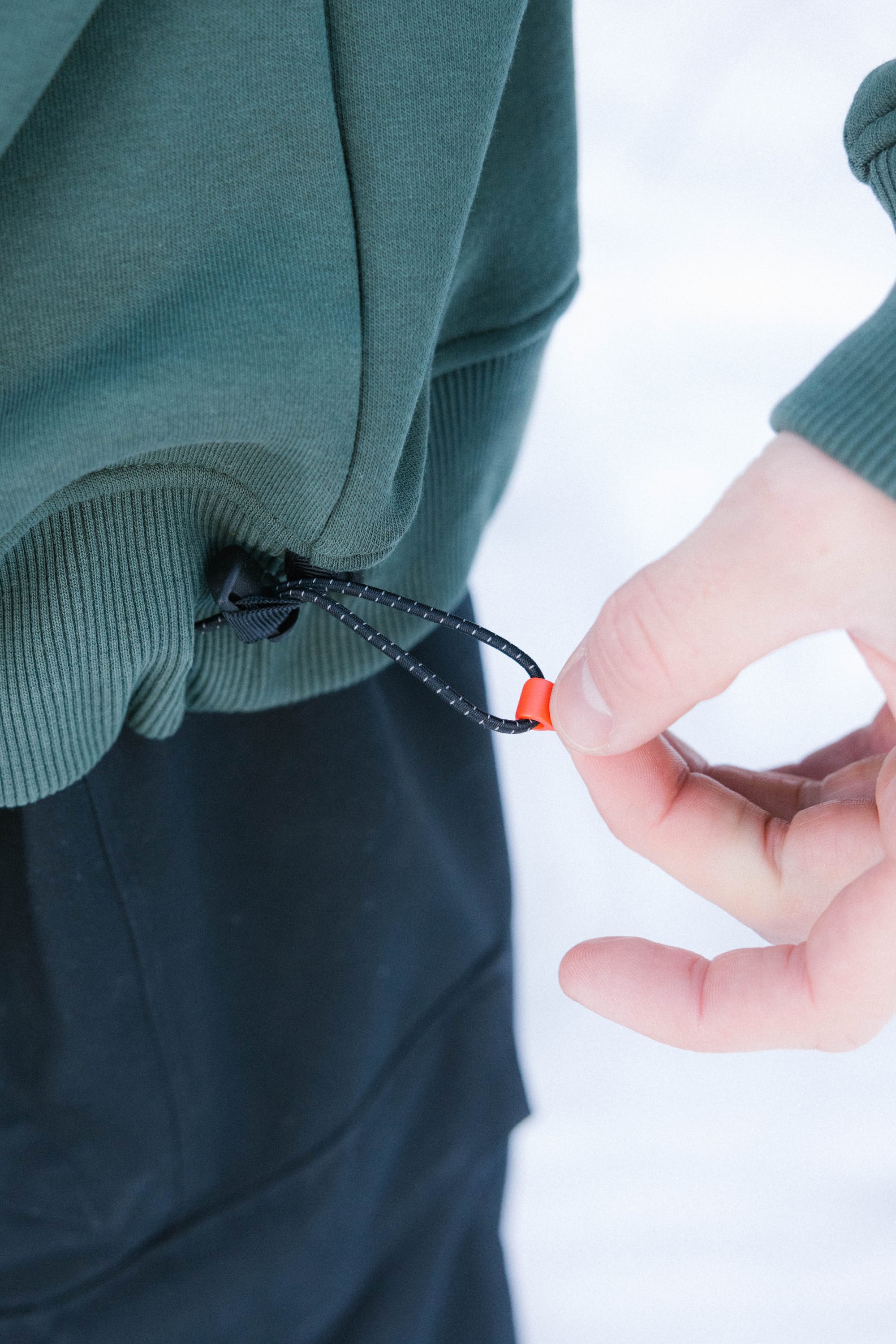 Someone adjusts the hem drawstring of a green 686 Knockout Logo Premium Heavyweight Pullover Hoody, pulling a black cord with an orange toggle against a softly blurred, light background.