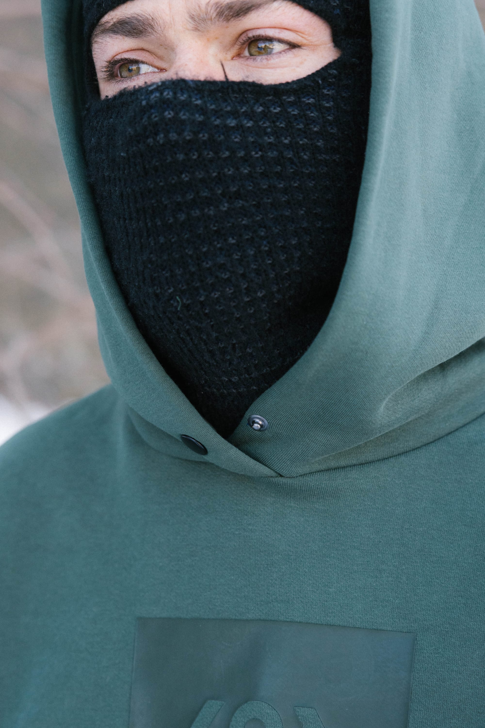 Someone stands outdoors in a cold setting, visible only from the eyes up, wearing a black knit face mask and the 686 Knockout Logo Premium Heavyweight Pullover Hoody by 686. The blurred background emphasizes the chilly atmosphere.