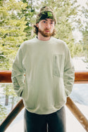 A bearded man with long hair, wearing a green beanie and the 686 Mens HUF Fleece Crewneck, stands outside on a wooden balcony with snowy trees in the background.
