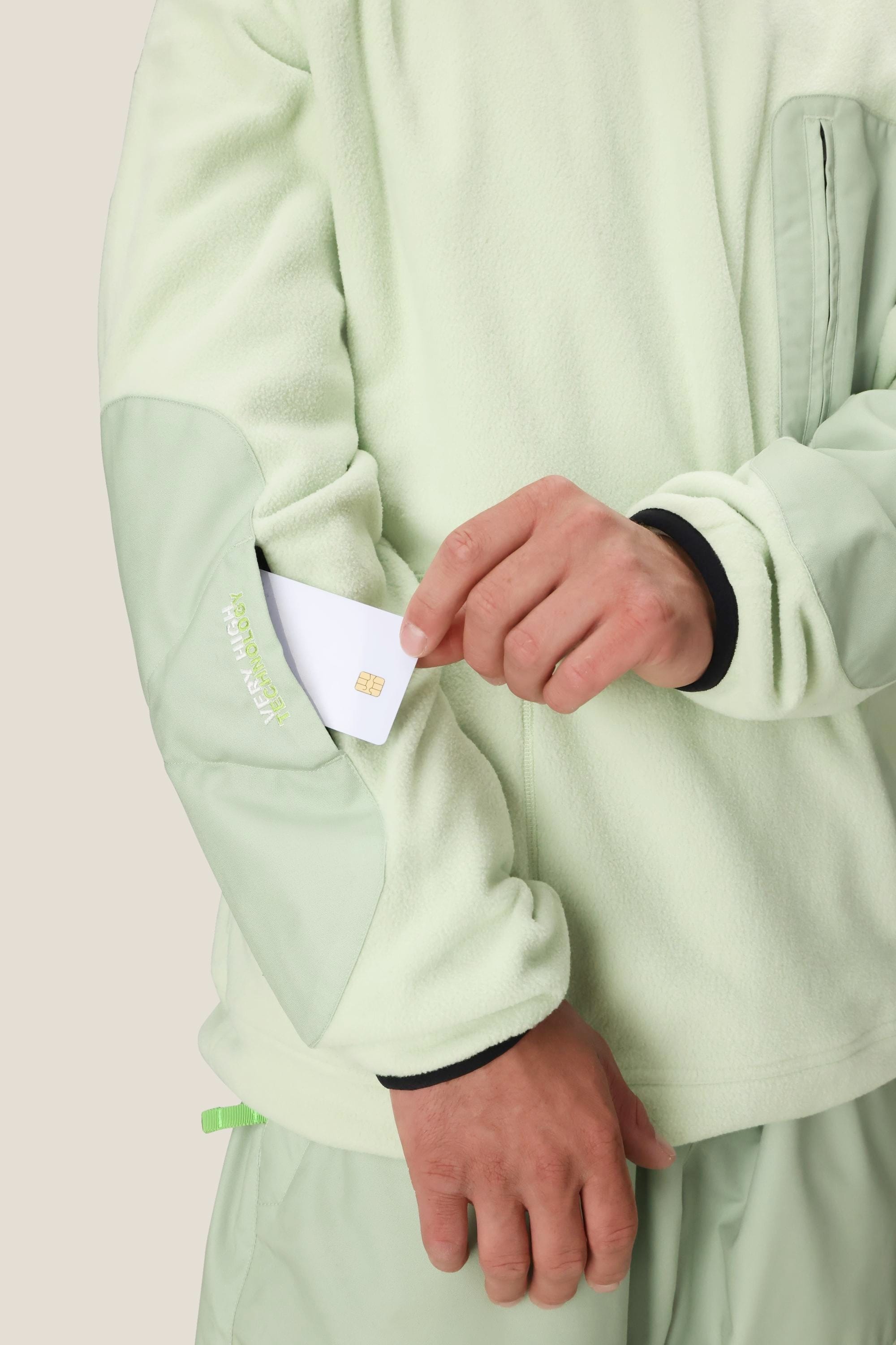 A person wearing the 686 Mens HUF Fleece Crewneck in light green places a white chip card into a zippered sleeve pocket on their right arm. Only the torso and arms are visible.