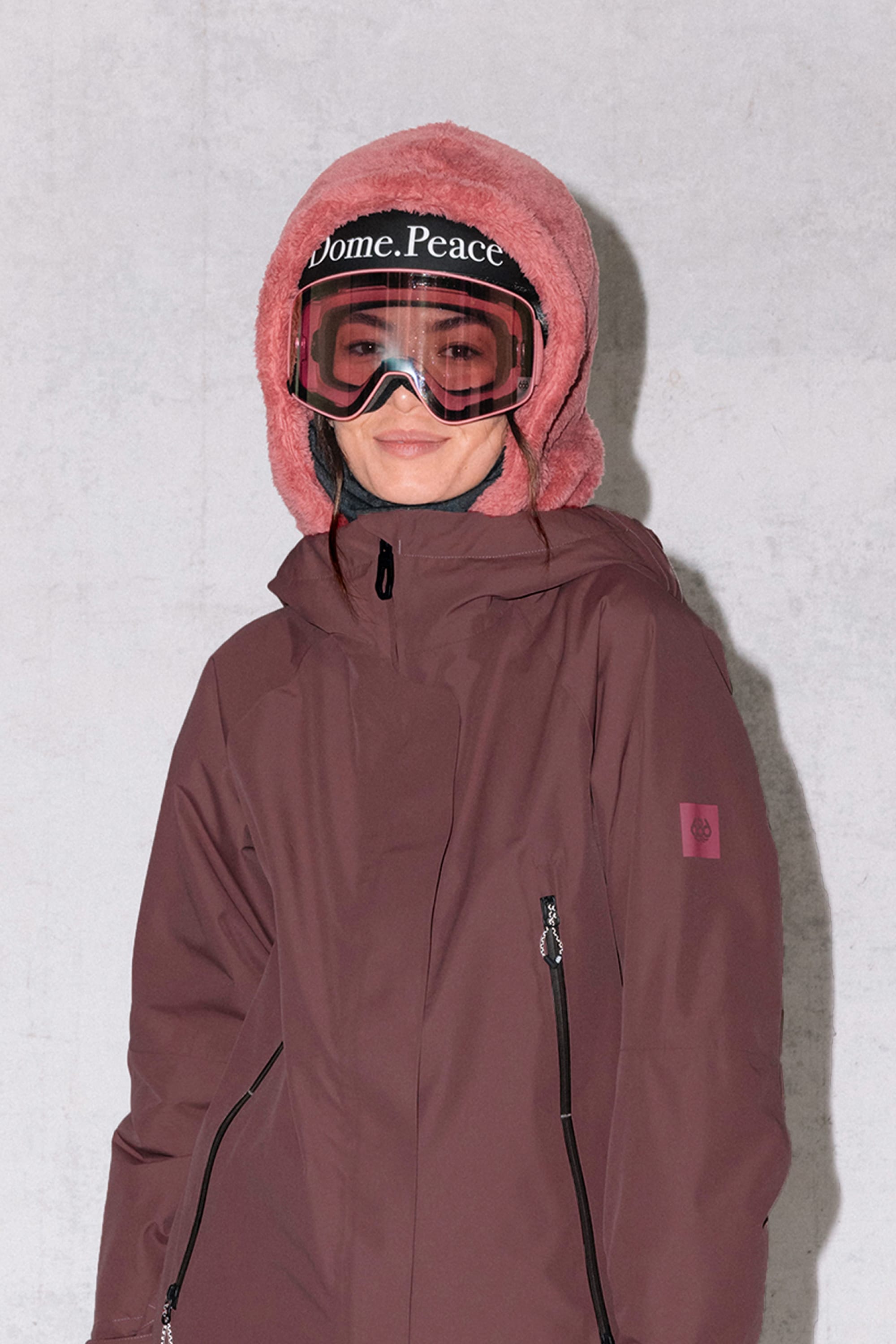 A person in a maroon winter jacket and pink fuzzy hat stands by a light gray wall, smiling, with the 686 Tarmac Sherpa Hood by 686—a polyester knit hood—visible under the hat, and wearing ski goggles labeled Dome. Peace.
