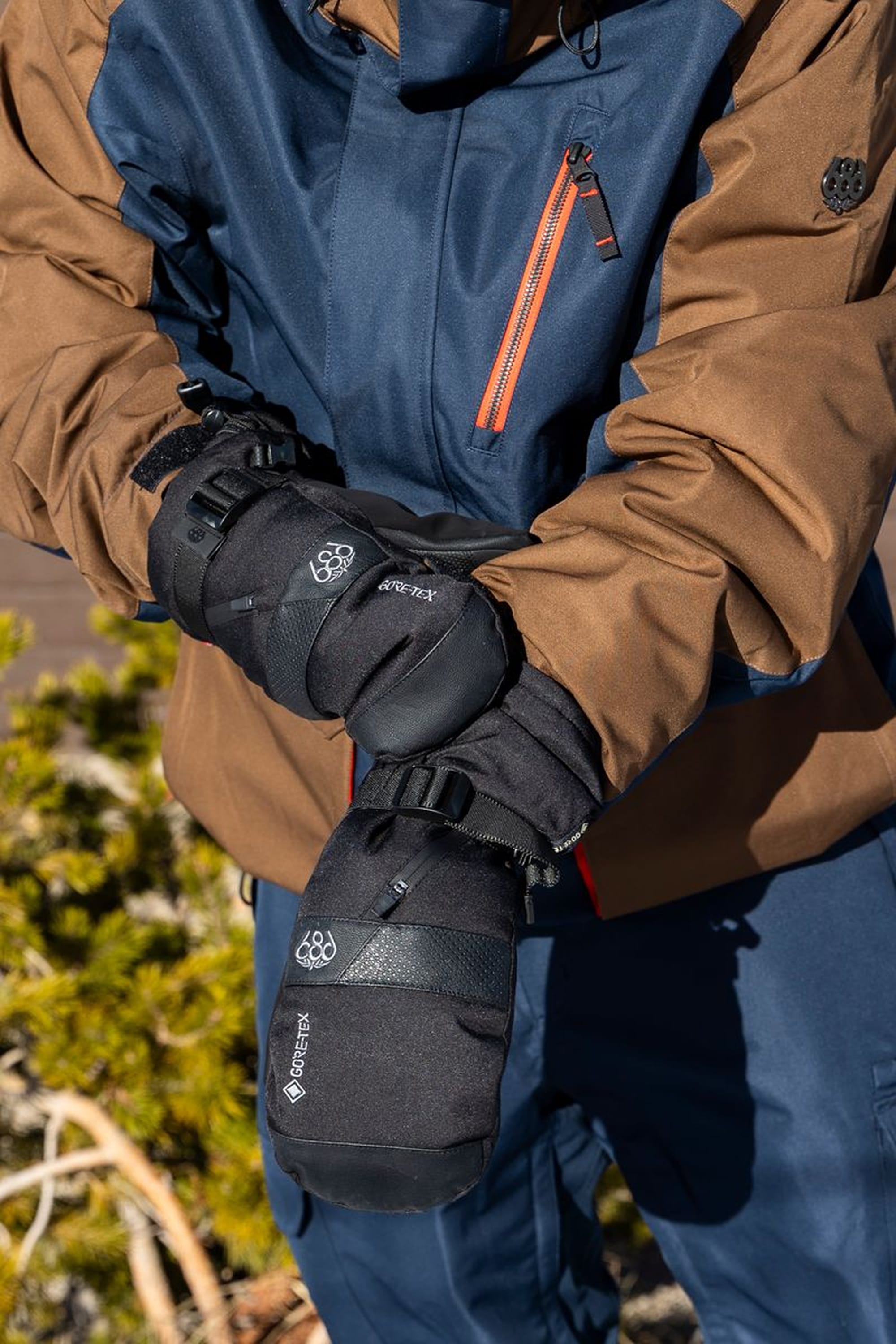 Wearing a brown and navy jacket, a person fastens the wrist strap on their black 686 Men’s GORE-TEX® SMARTY® 3-In-1 Gauntlet Mitt outdoors, with greenery blurred in the background.