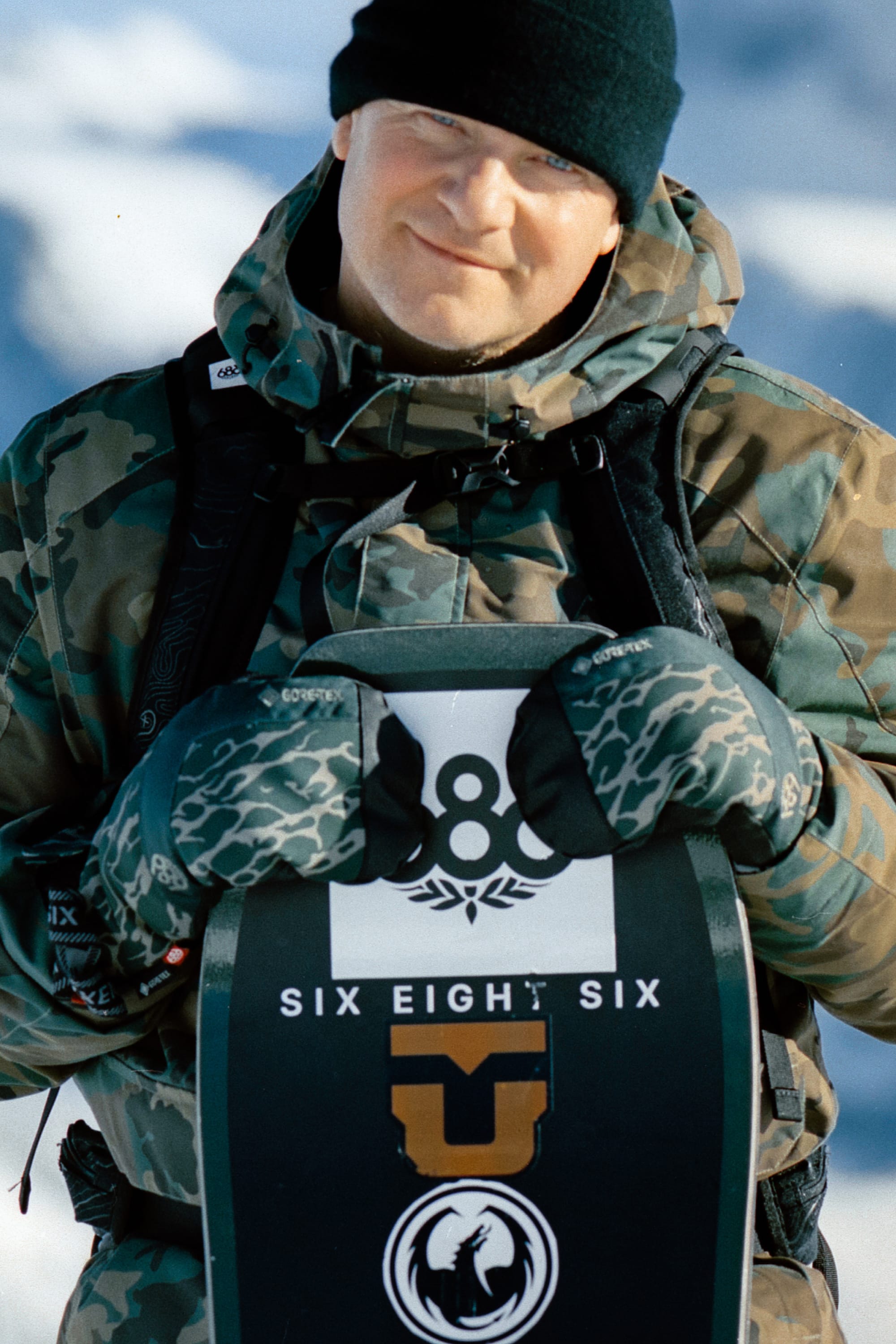 A person in camo winter gear and 686 Men's GORE-TEX Linear™ Under Cuff Mitts holds a snowboard with 686 logos, smiling outside in the snow and wearing a black beanie.