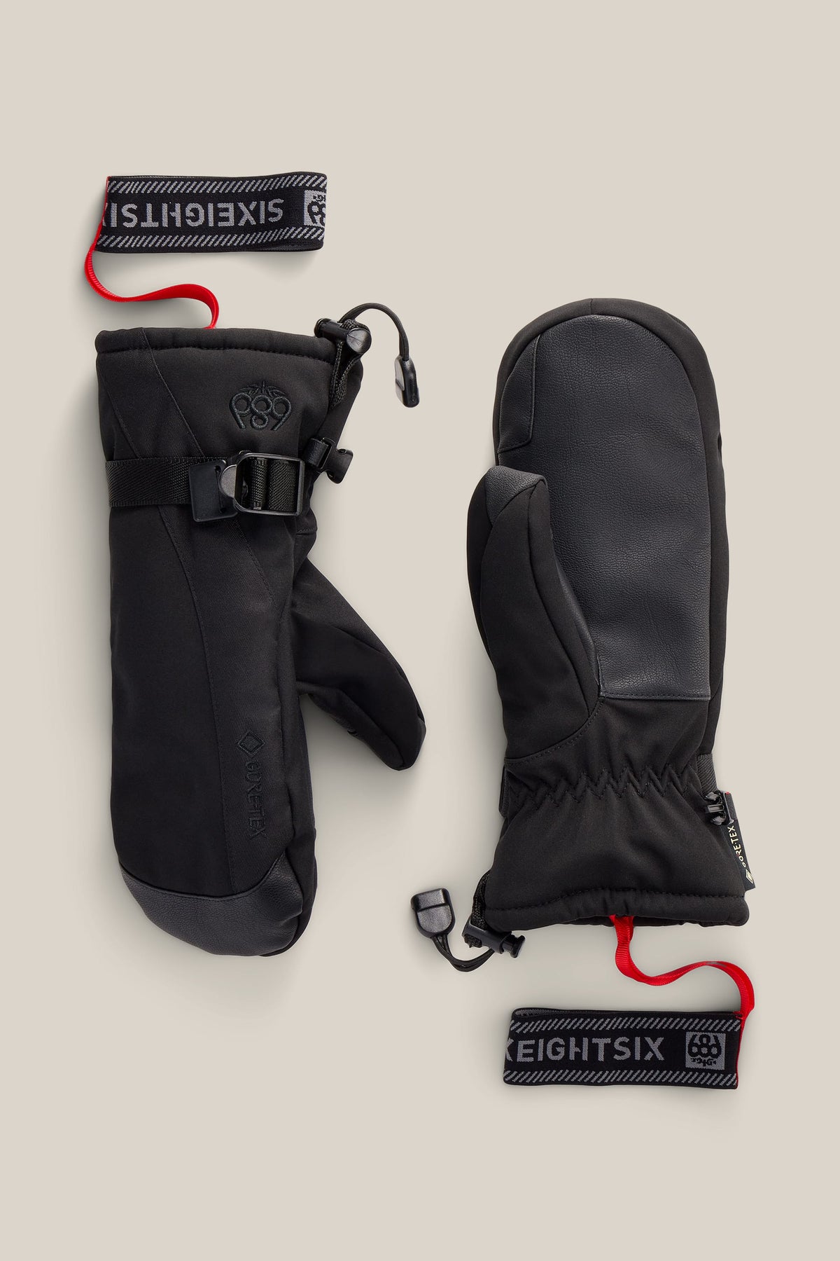A pair of 686 Women’s GORE-TEX® Linear™ Mitts in black, featuring insulated construction with wrist straps, drawcords, leather palms, and red leash attachments, displayed flat on a neutral background.