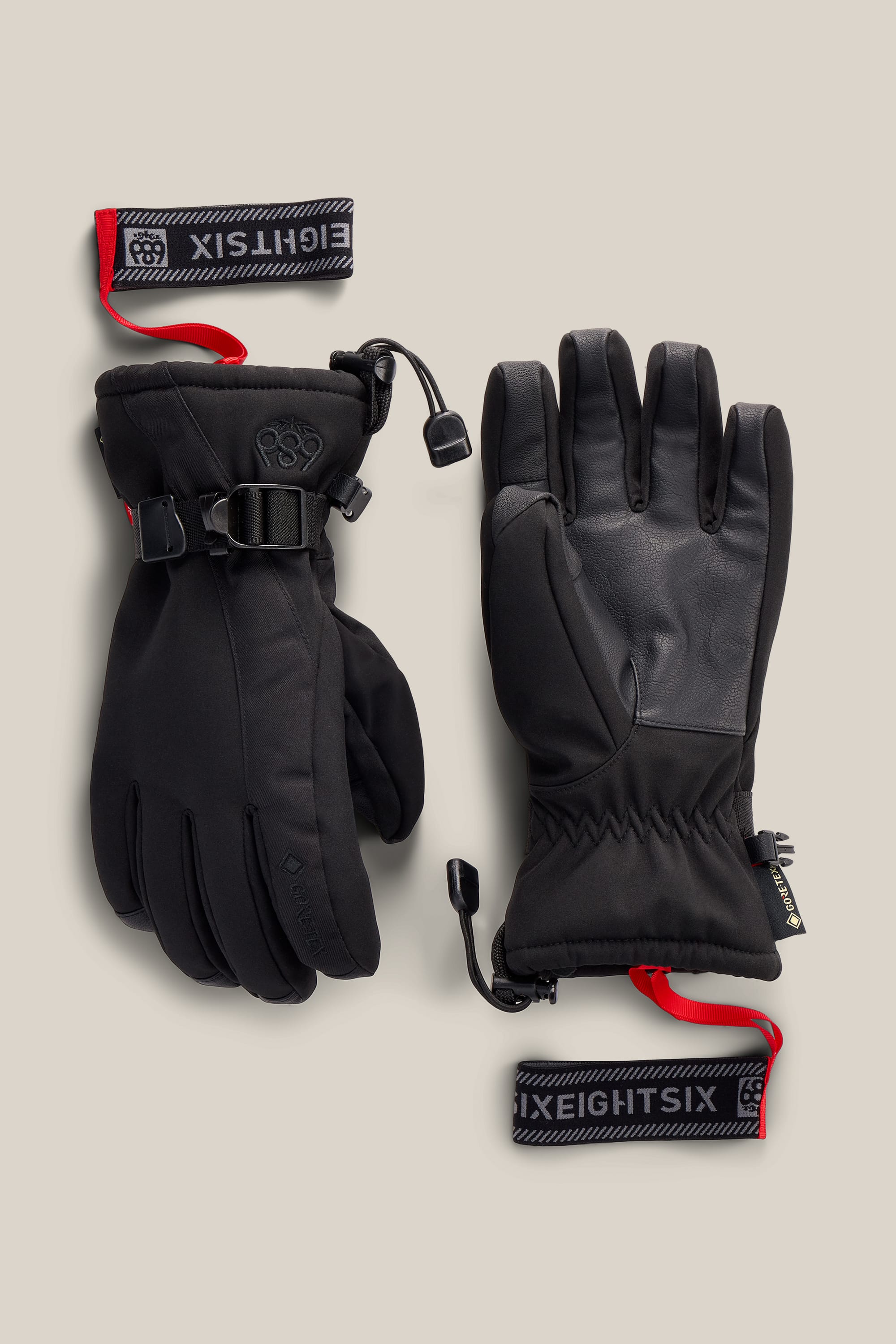 A pair of 686 Women’s GORE-TEX® Linear™ Gloves in black, featuring wrist straps and red pull tabs labeled “686.” One glove is shown with the palm side visible, while the other displays the back side with a buckle.