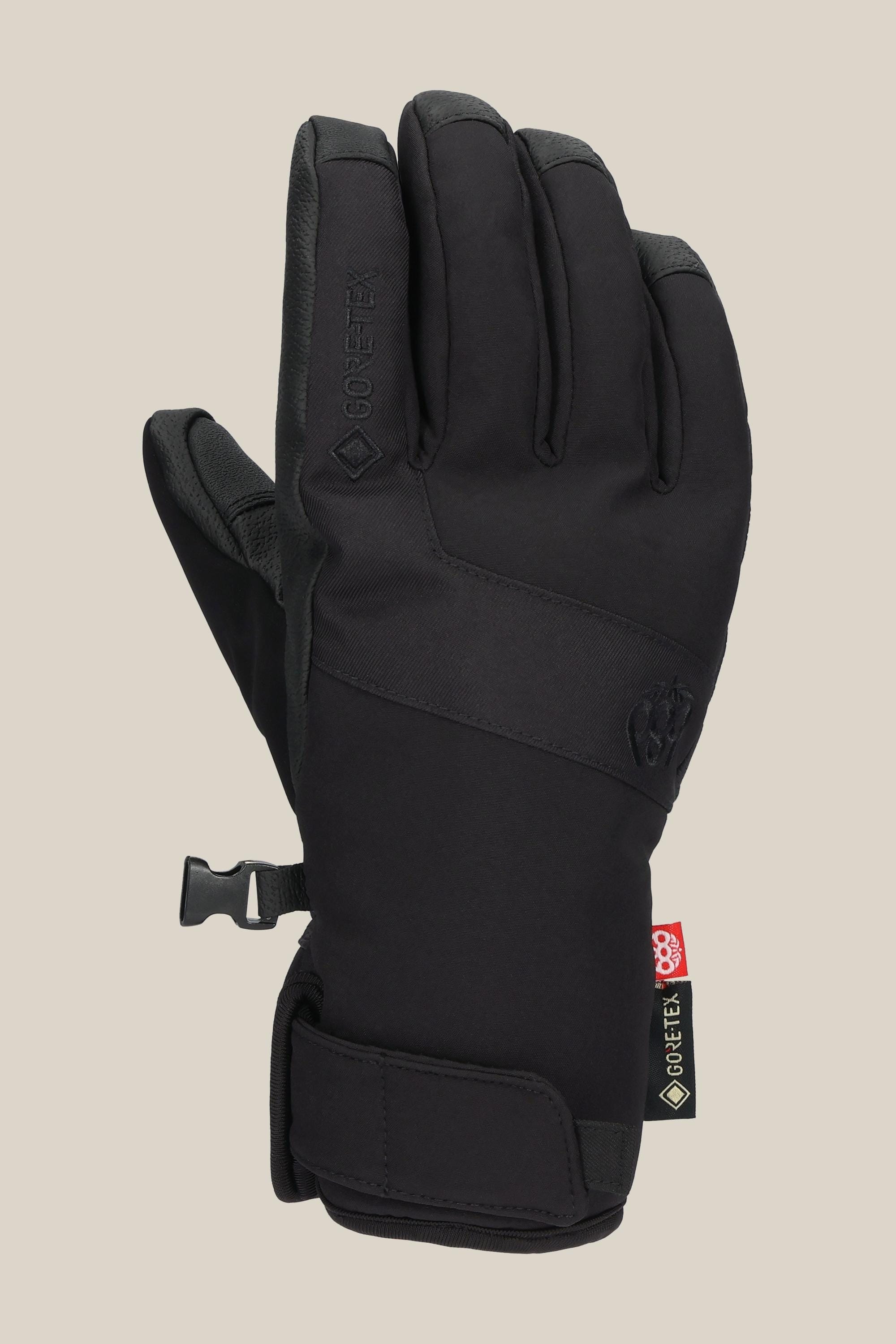 The 686 Women's GORE-TEX Linear™ Under Cuff Glove is shown upright, palm away, in black with a Velcro wrist strap, GORE-TEX tag, and side clip on a beige background—a stylish winter glove from 686.