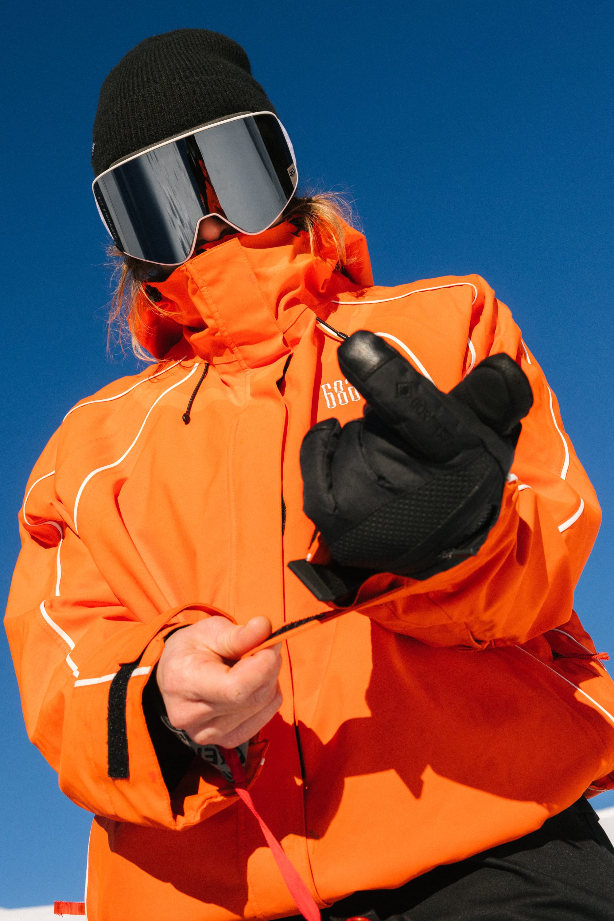 Wearing an orange winter jacket, a pair of 686 Women's GORE-TEX SMARTY® 3-In-1 Gauntlet Gloves, and ski goggles, a person stands outdoors against a blue sky, adjusting their strap. Their face is mostly hidden by the jacket, hat, and goggles.