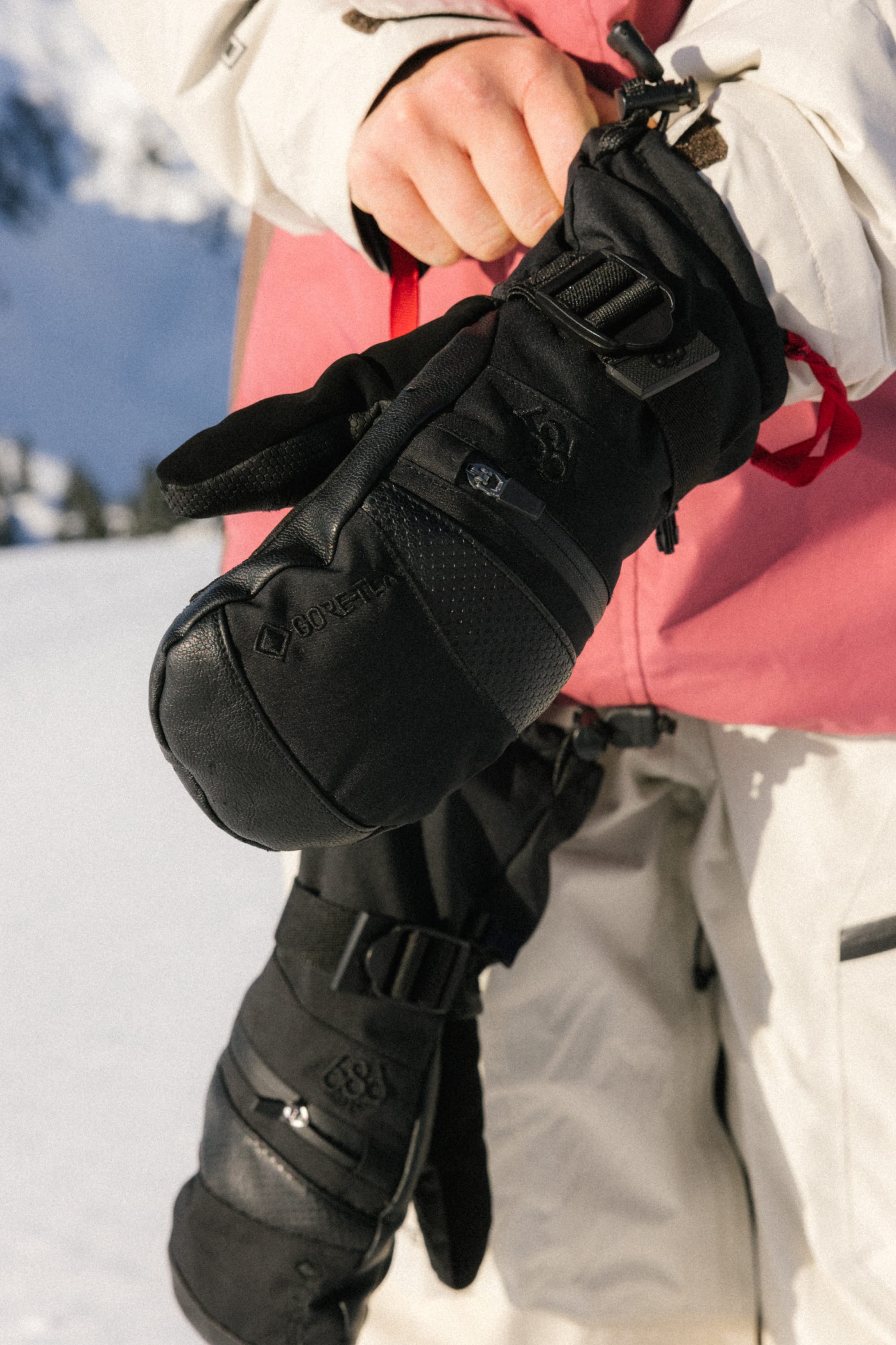 Outdoors with snowy mountains in the background, a person in a white and pink winter jacket holds 686 Women’s GORE-TEX SMARTY® 3-In-1 Gauntlet Mitts by 686 in one hand.