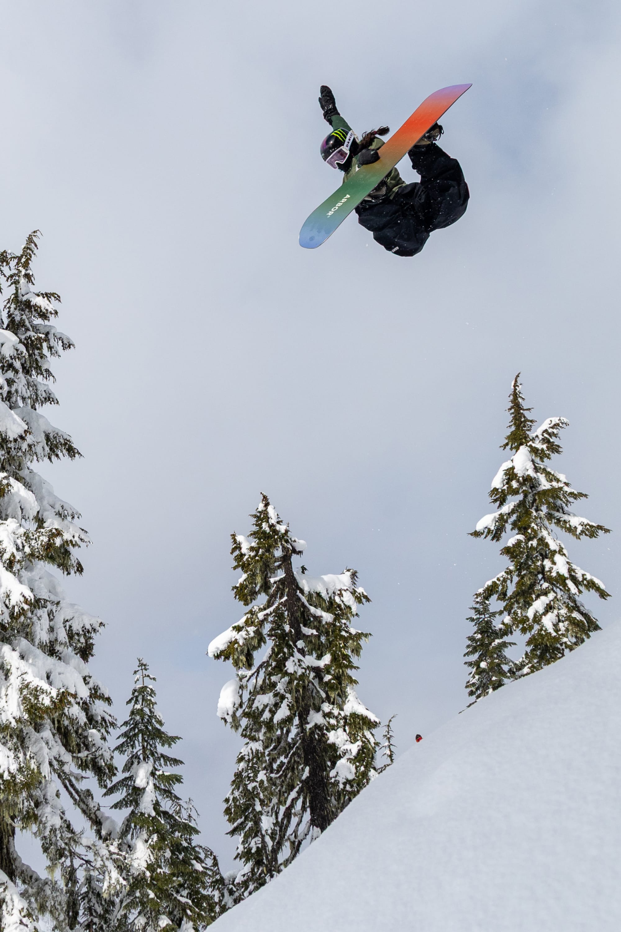 Wearing the 686 Women’s GORE-TEX® SMARTY® 3-In-1 Gauntlet Mitt from 686, a snowboarder performs a high jump trick above a snowy slope, surrounded by tall pine trees blanketed in snow on a cloudy winter day.