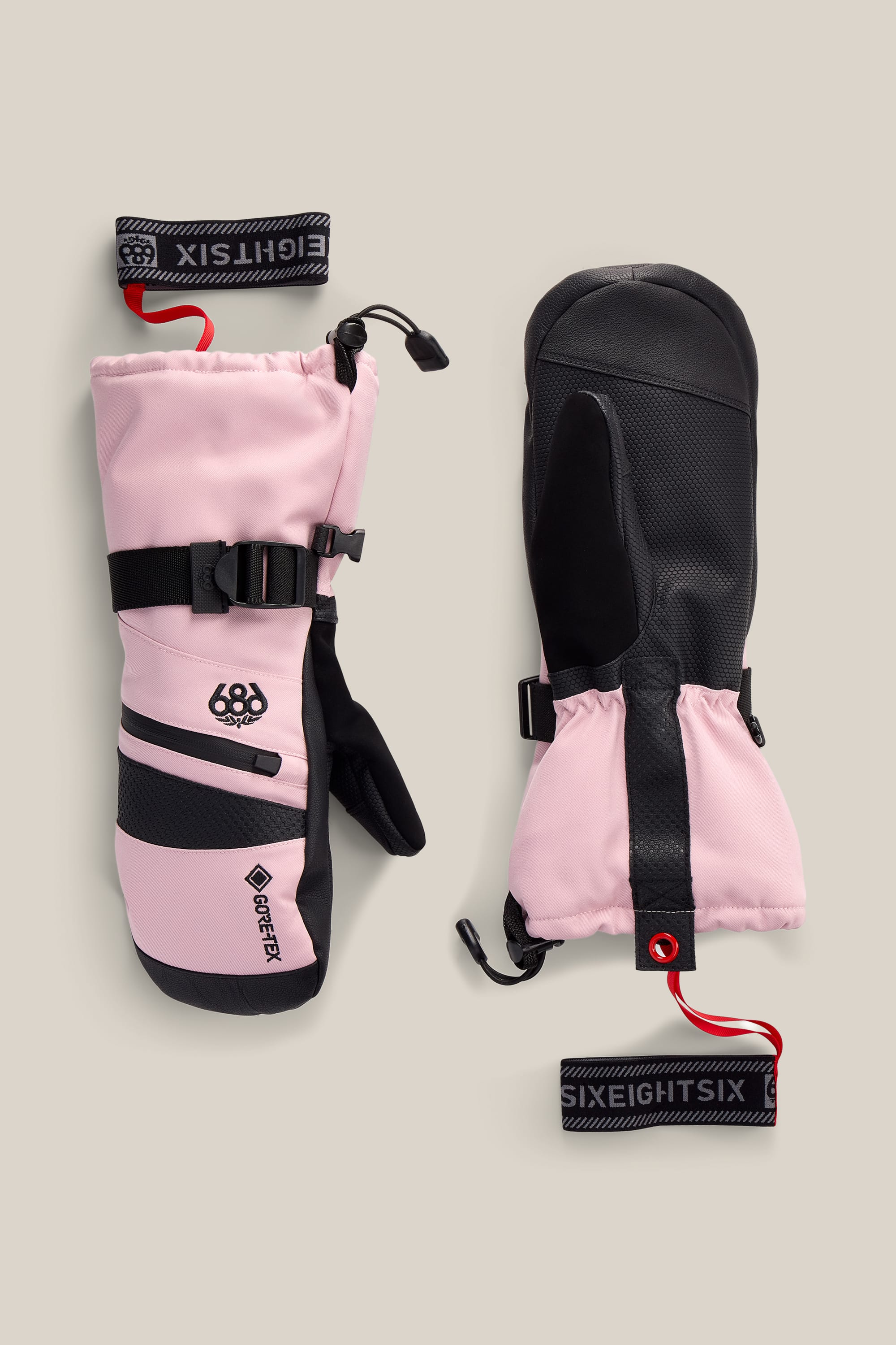 A pair of 686 Women’s GORE-TEX® SMARTY® 3-In-1 Gauntlet Mitts in pink and black, featuring adjustable wrist straps with “SIXEIGHT SIX” printed in white on a black and red background and black palms. The mittens are displayed on a beige surface.