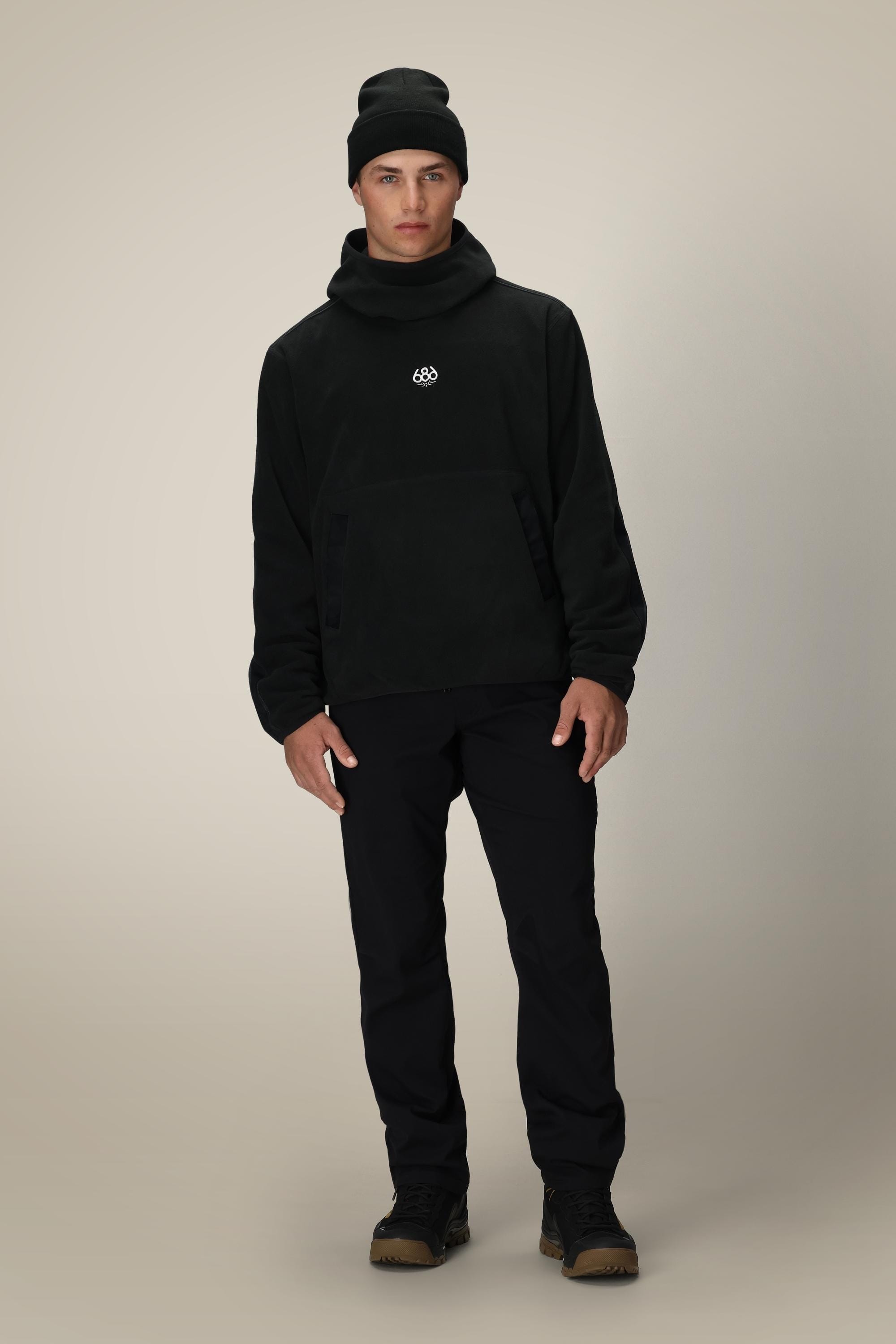 A young man stands facing forward, wearing the 686 Mens Clava Fleece Hoody in black with a small white logo, paired with black pants and brown shoes, set against a plain light beige background.