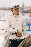 A young man wearing the 686 Mens Clava Fleece Hoody and a cap with sunglasses on top leans against a boat railing, holding a phone. Water, blurred boats, and overcast skies form the background.