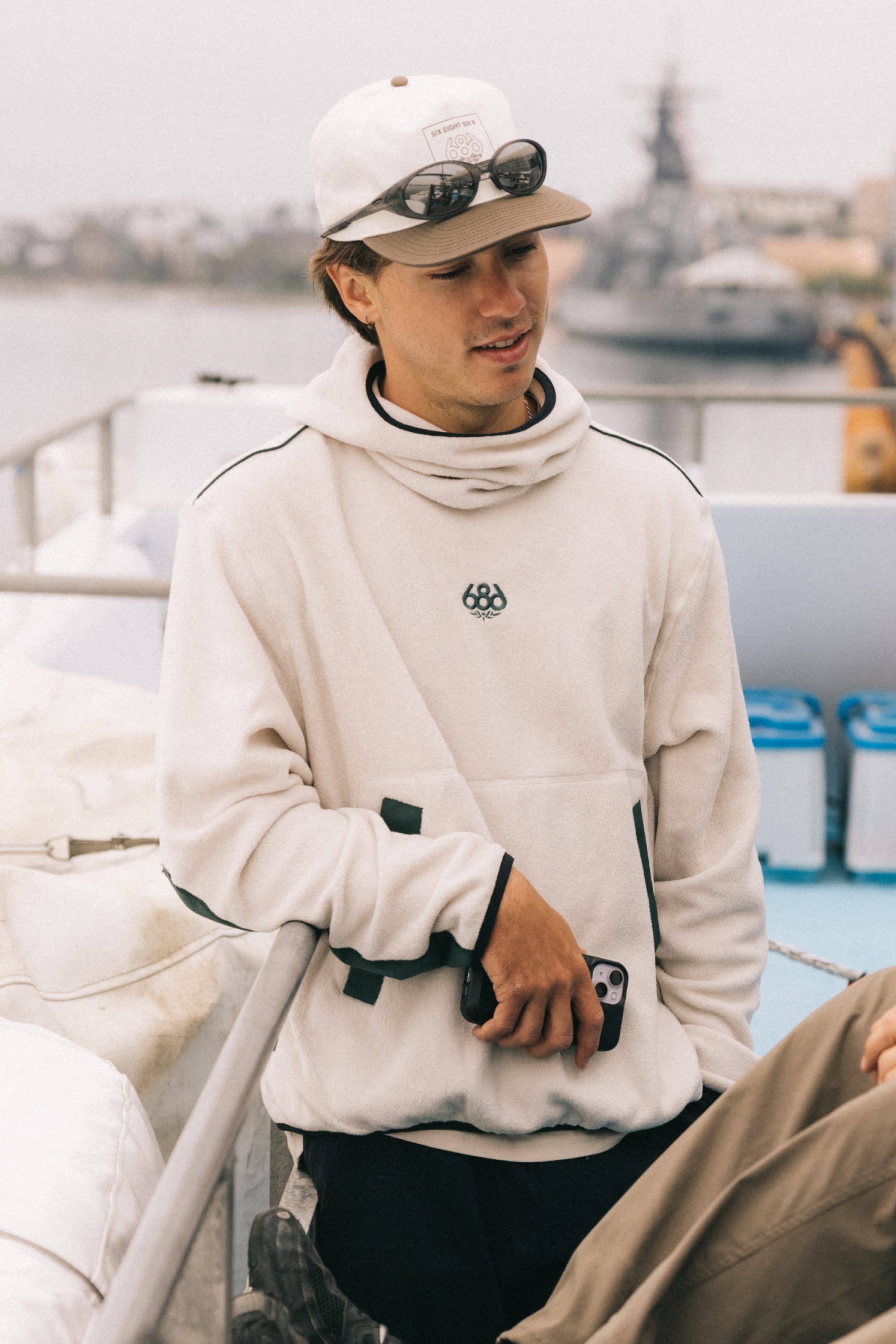 A young man wearing the 686 Mens Clava Fleece Hoody and a cap with sunglasses on top leans against a boat railing, holding a phone. Water, blurred boats, and overcast skies form the background.
