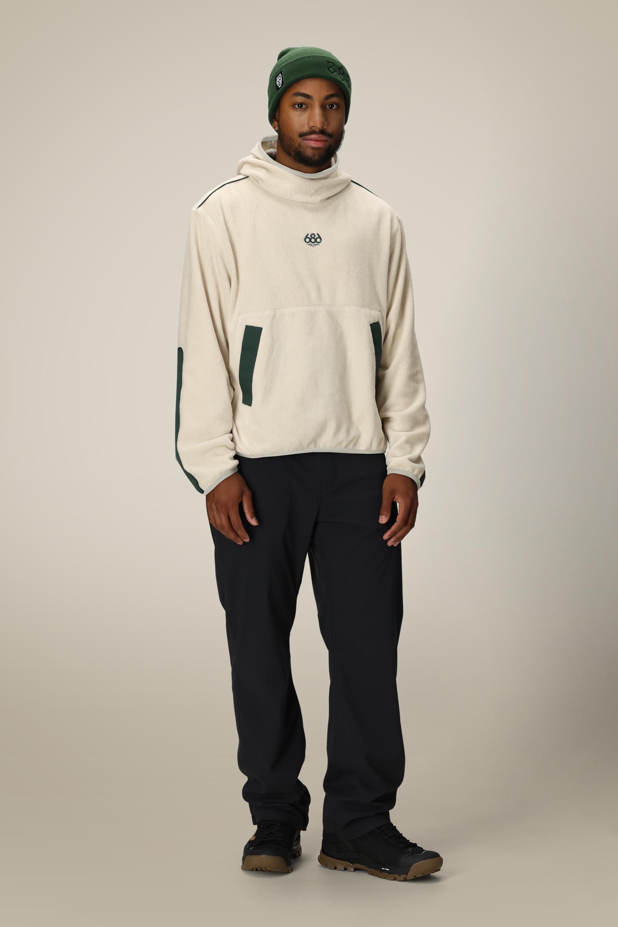 A person wearing the 686 Mens Clava Fleece Hoody in cream with dark green accents, paired with dark pants, brown shoes, and a green beanie, stands facing the camera against a plain light background.