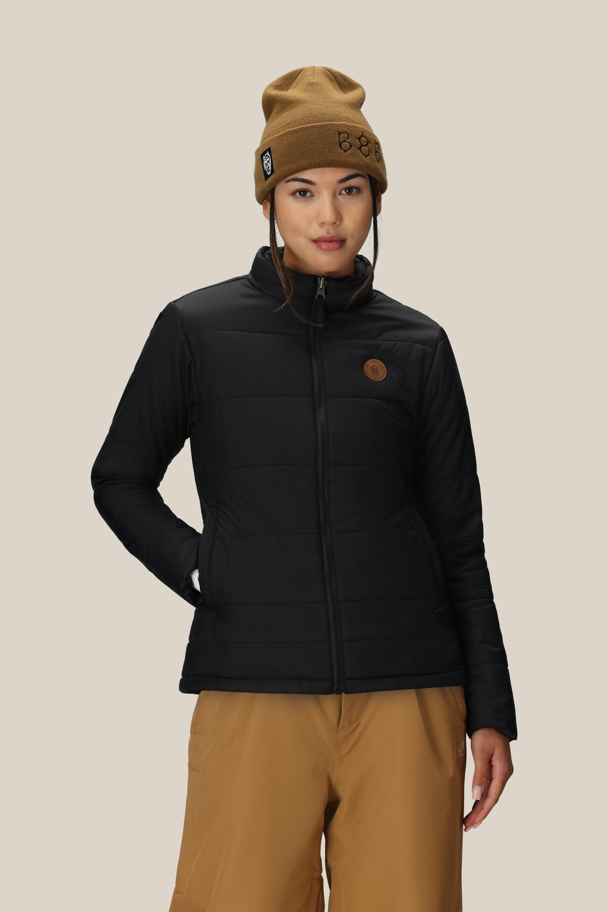 A person wearing the 686 Women’s Thermal Puff Insulated Jacket from 686, paired with a tan beanie and tan pants, stands against a plain beige background looking slightly to the side with a neutral expression.