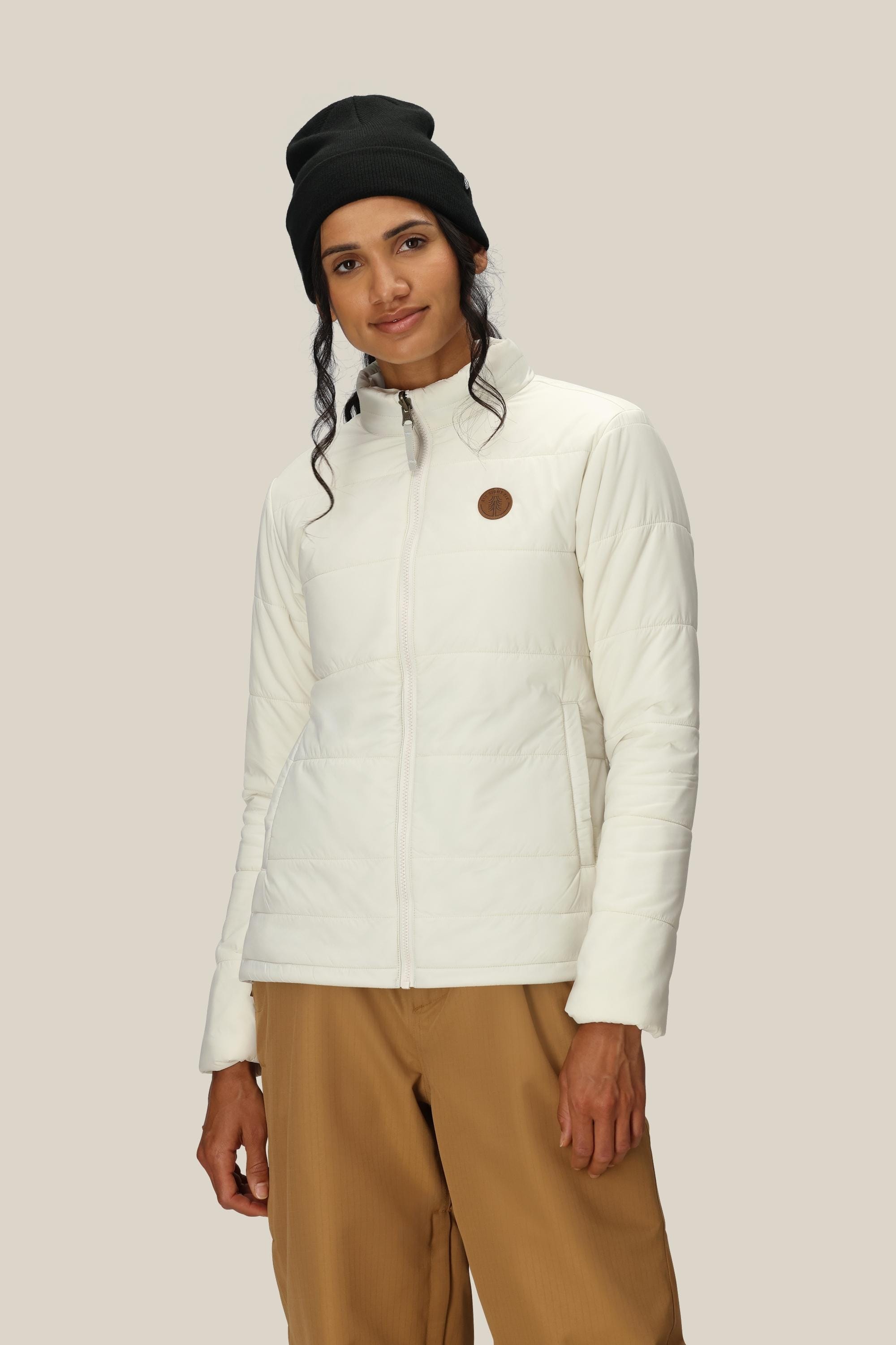 A person wearing the 686 Women’s Thermal Puff Insulated Jacket in white, paired with brown pants and a black beanie, stands against a plain beige background, looking at the camera with a slight smile.