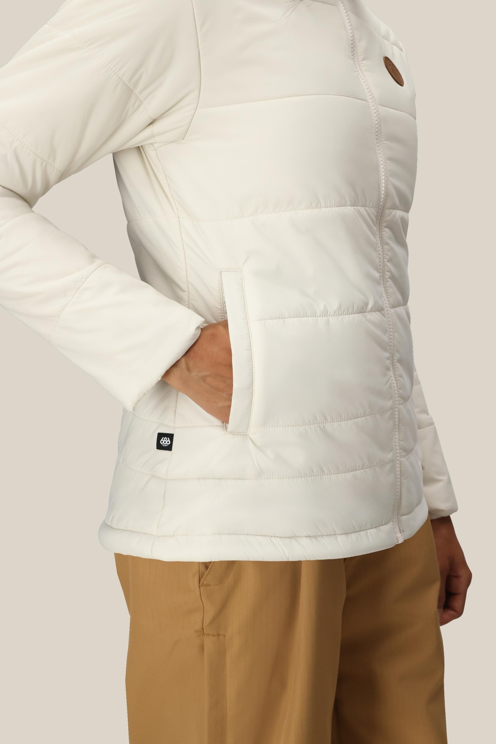 A person wearing the 686 Womens Thermal Puff Insulated Jacket with their right hand in a side pocket, paired with tan pants, stands against a plain beige background.