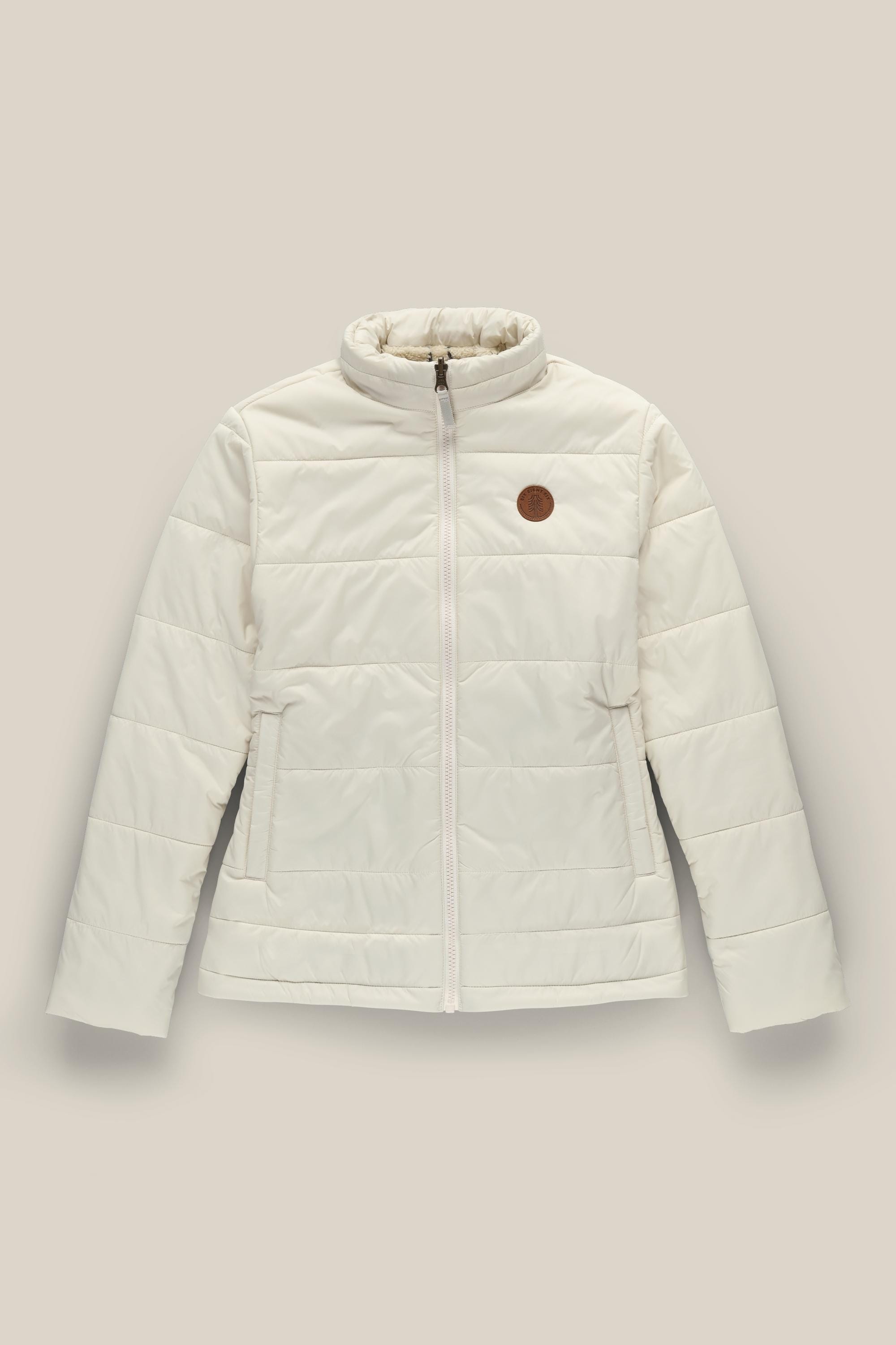 The 686 Womens Thermal Puff Insulated Jacket by 686 is a polyester puffer with a front zipper, side pockets, high collar, and a round brown patch on the left chest. Shown against a plain beige background.