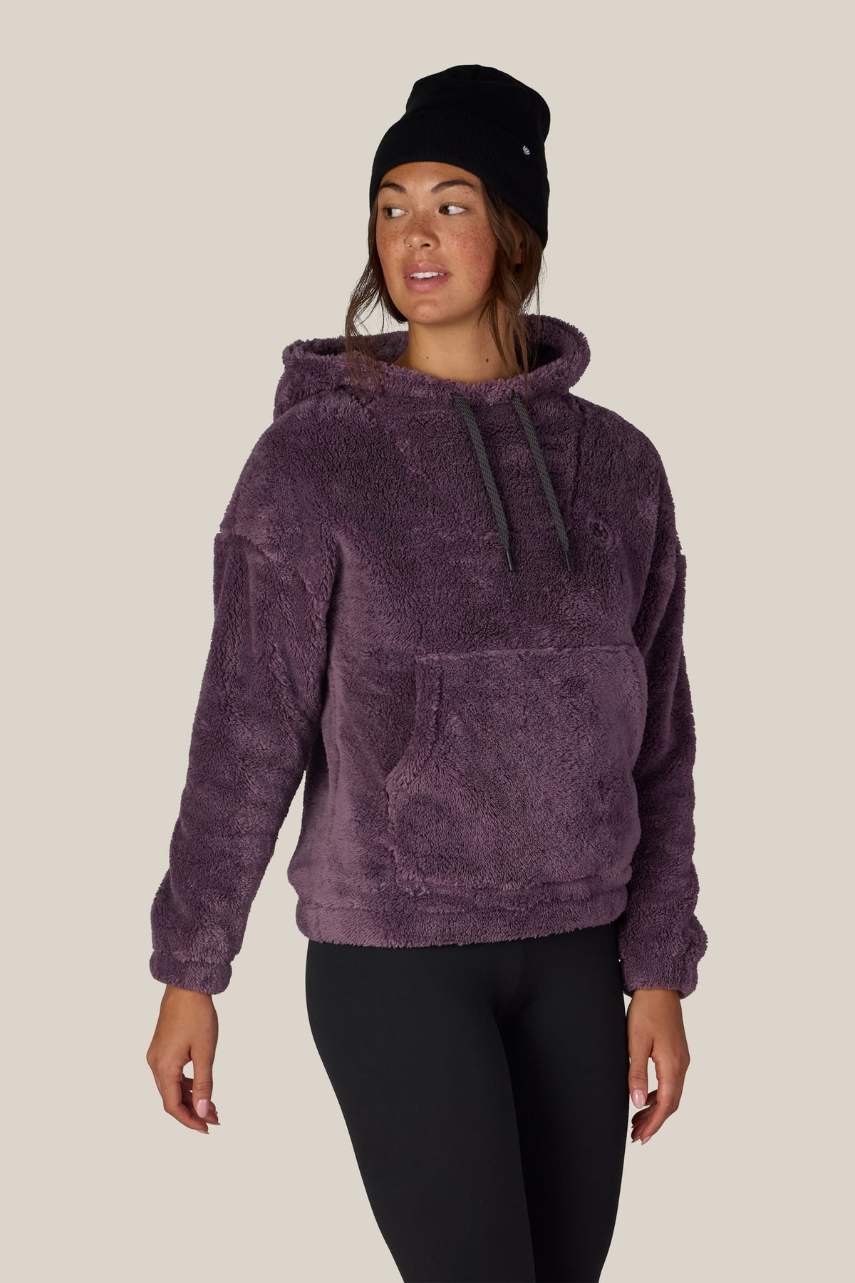 A person wearing the 686 Women’s Sherpa Hoody in purple by 686, paired with a black knit beanie and black leggings, stands against a plain beige background, looking to the side.