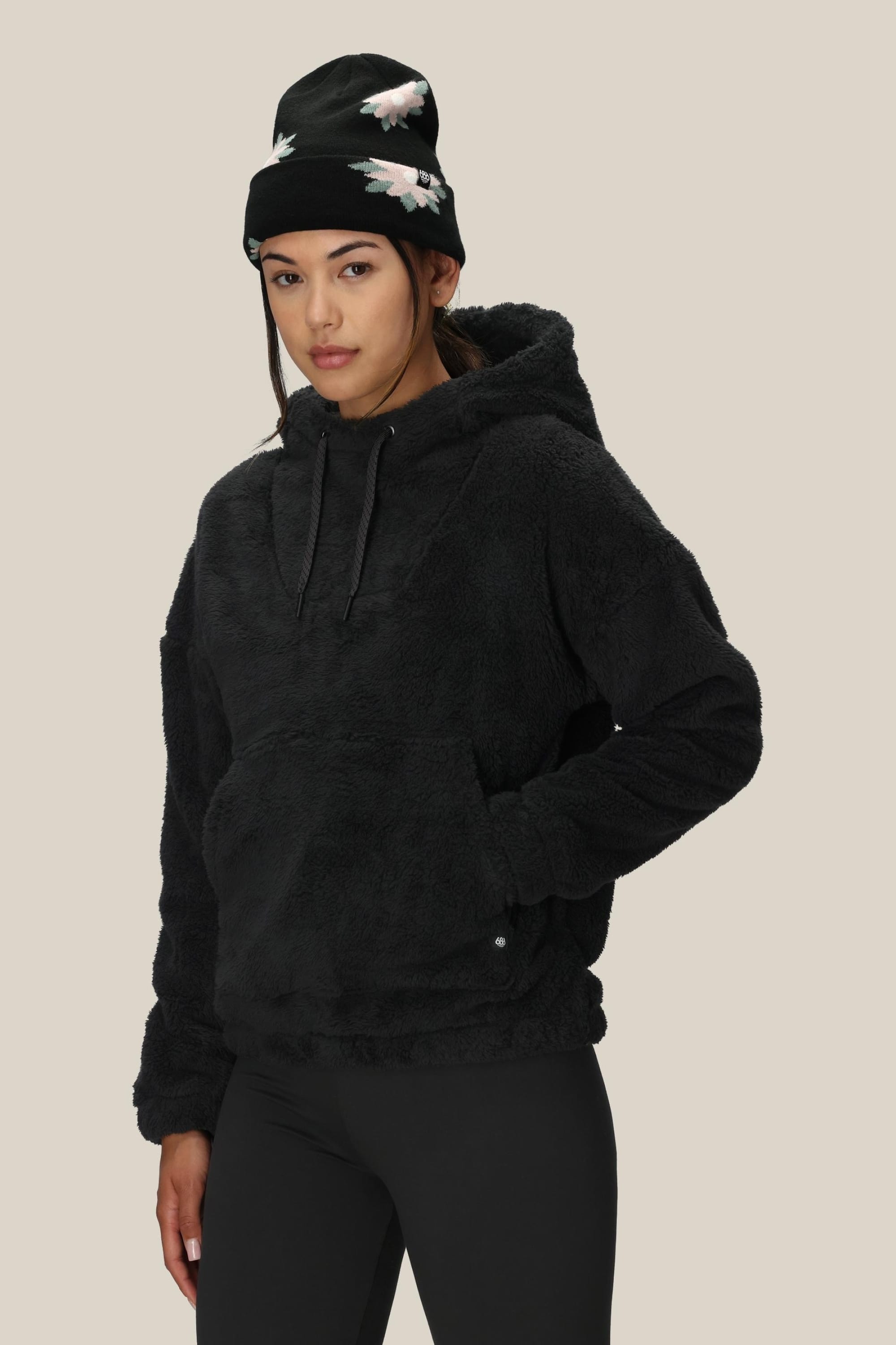 A woman wears the 686 Womens Sherpa Hoody—a fuzzy black polyester jacket by 686—paired with black leggings and a beanie with white floral patterns, standing against a plain light background and looking slightly to the side.