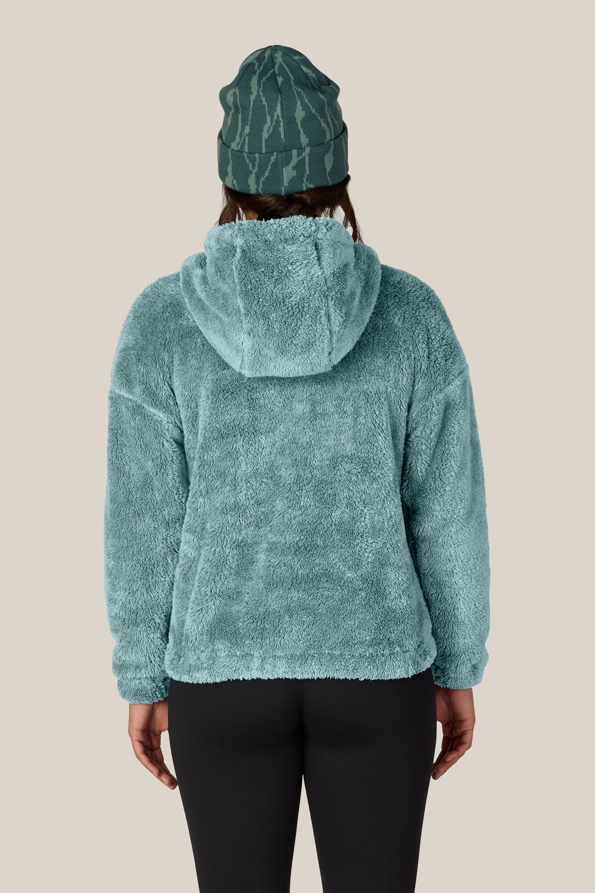 A person shown from the back against a plain light beige background wears the 686 Women’s Sherpa Hoody in teal, paired with black leggings and a green patterned beanie.
