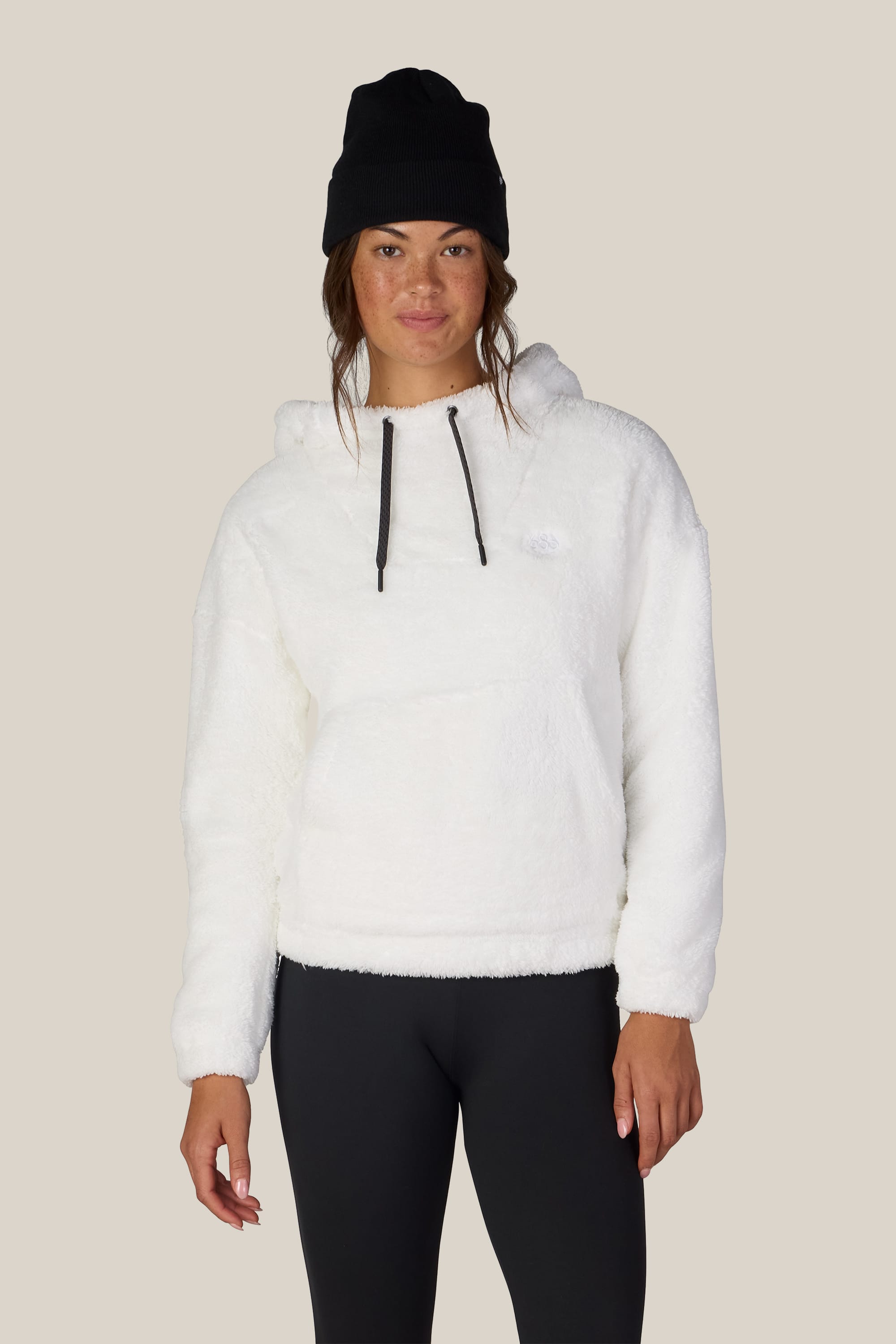 A woman wearing the 686 Women’s Sherpa Hoody in white, paired with black leggings and a black beanie, stands facing forward against a plain beige background.