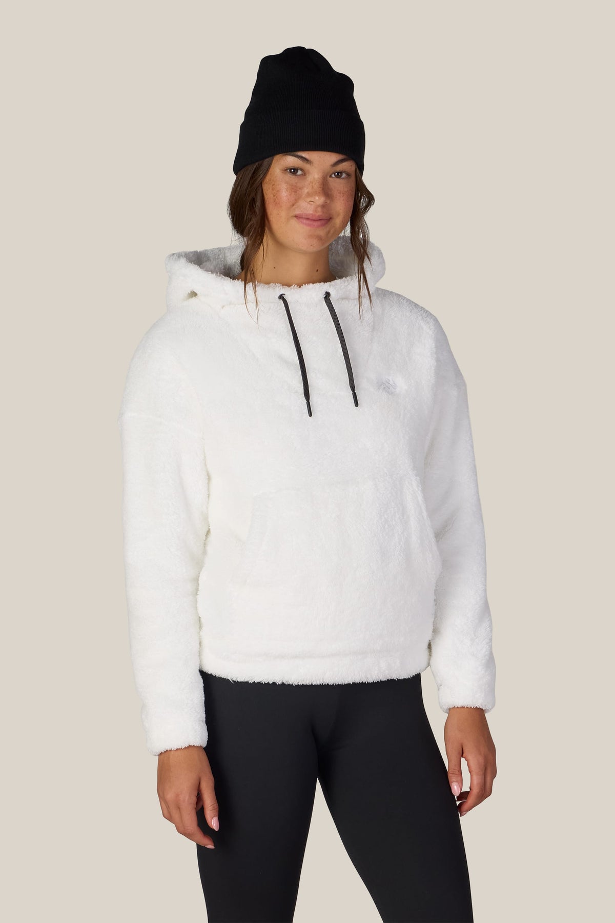 A woman stands facing forward, smiling slightly, against a plain beige background, wearing the 686 Women’s Sherpa Hoody in white by 686, along with a black beanie and black leggings.