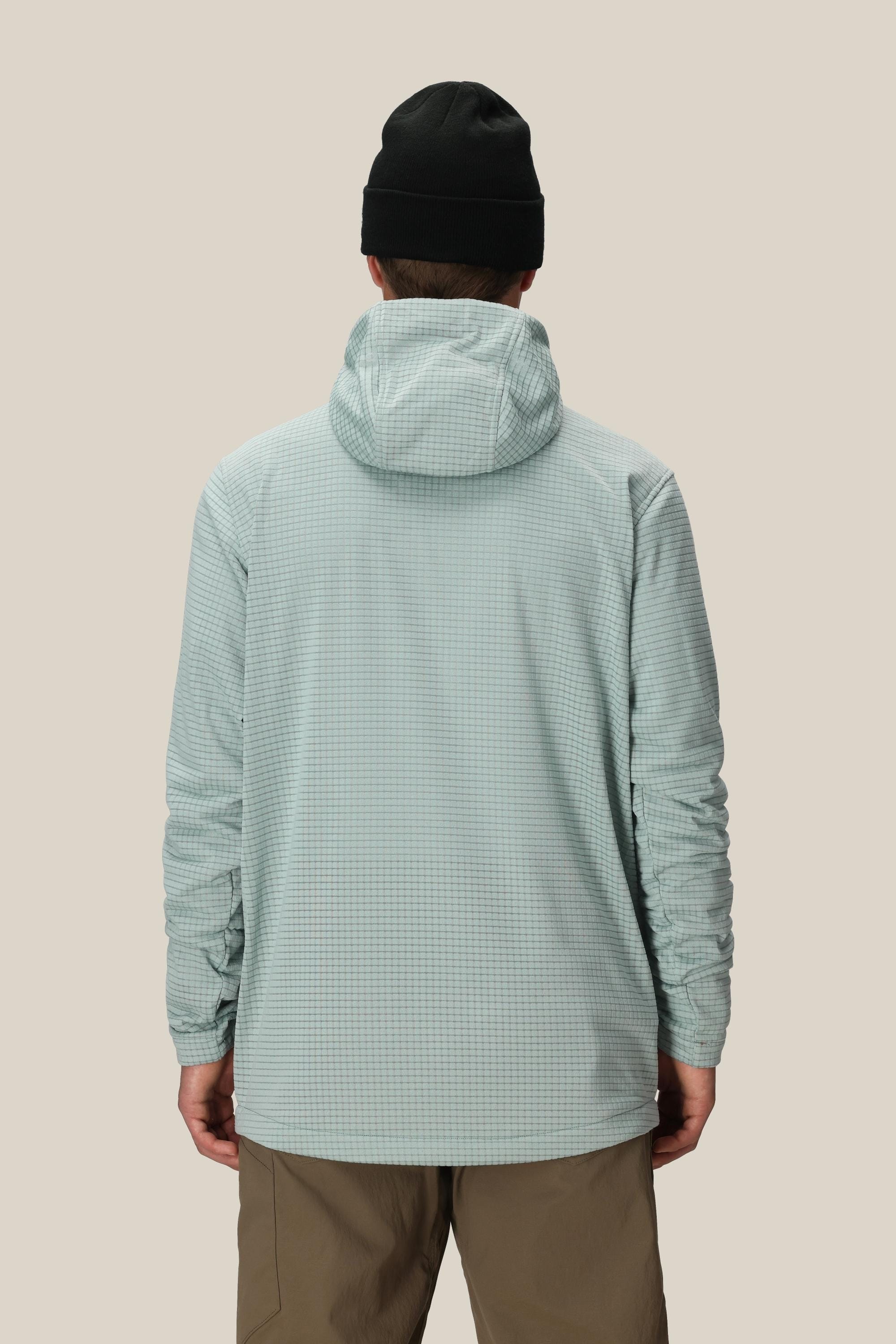 A person wearing a black beanie, khaki pants, and the 686 Men's Mission Grid™ Fleece Hoody by 686 in light blue stands with their back to the camera against a plain beige background.