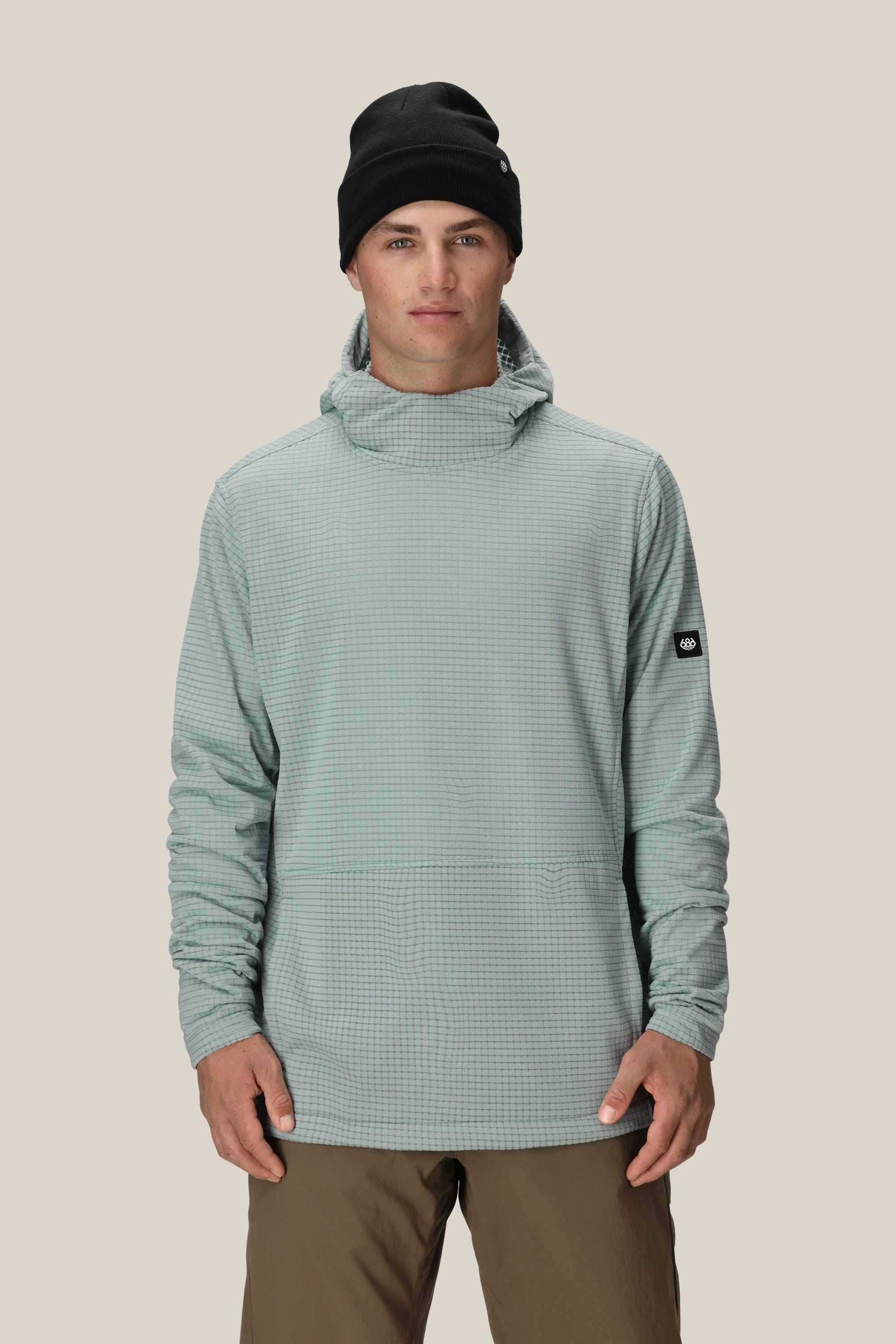 A man wears the 686 Men's Mission Grid™ Fleece Hoody in light gray, paired with a black beanie and brown pants, standing against a plain beige background.