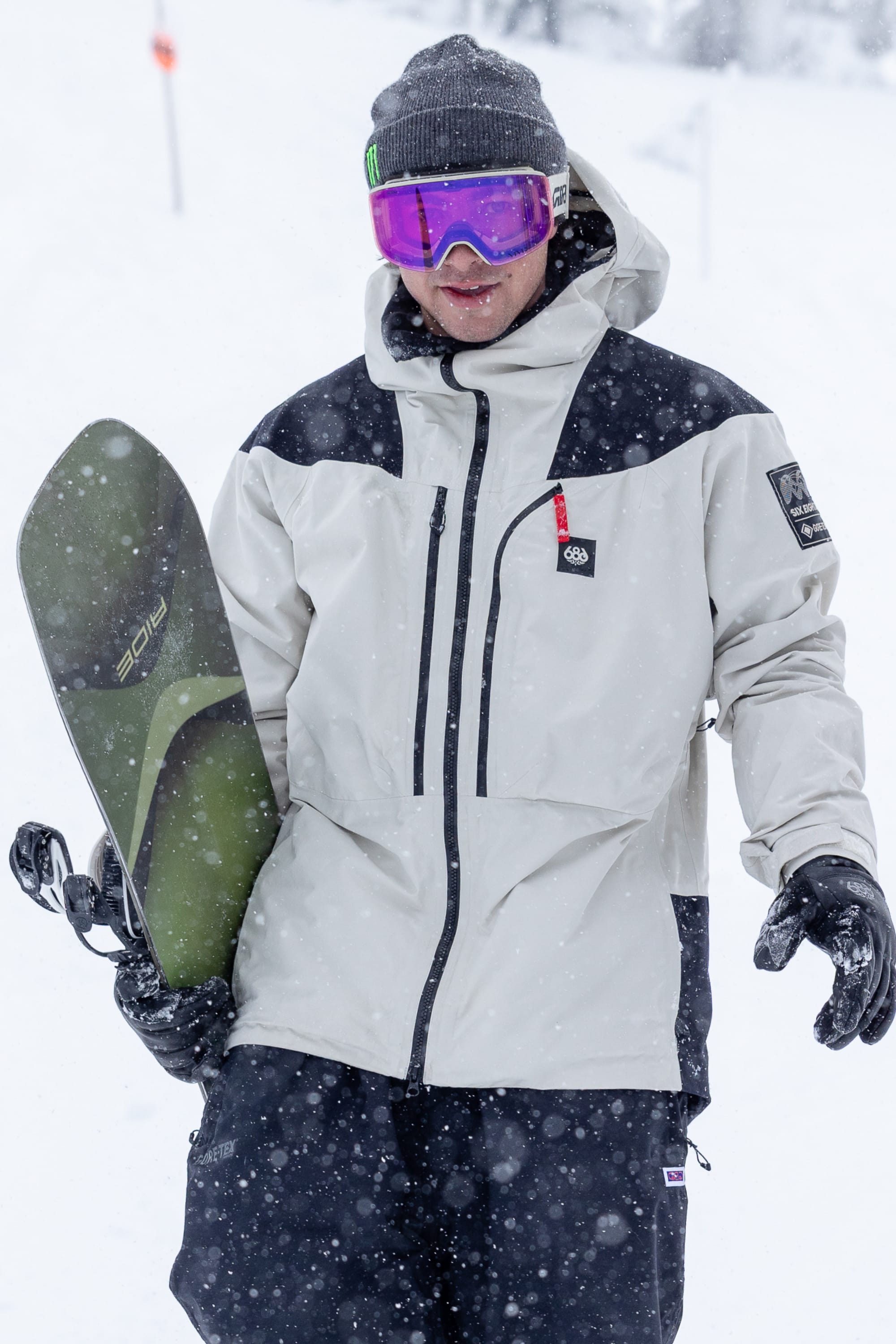 Wearing the 686 Men’s GORE-TEX® Dispatch™ Thermagraph® Jacket in light gray, along with black pants, a beanie, and pink ski goggles, a person holds a snowboard in a snowy outdoor setting as snow falls and the background is blurred.