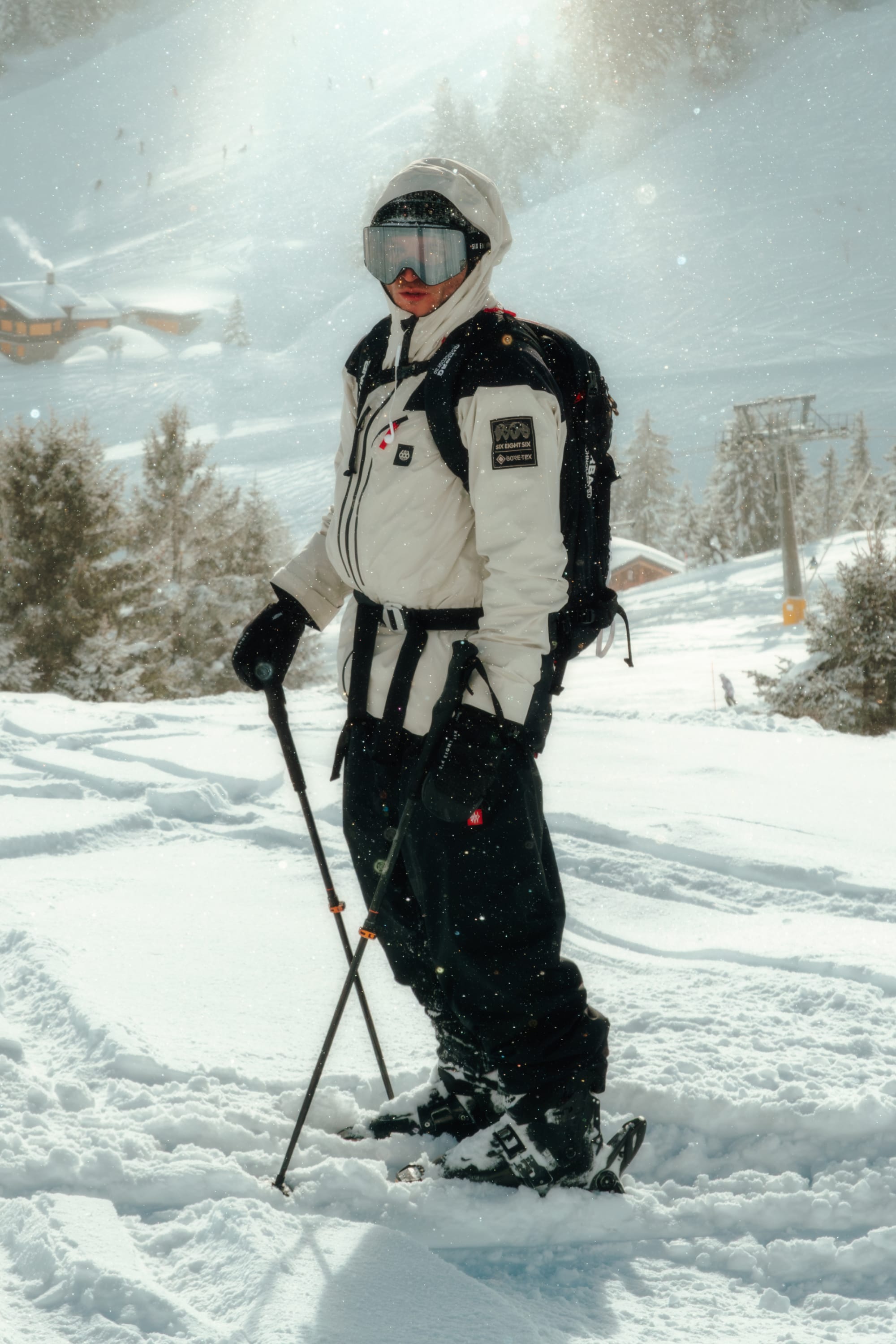 Wearing the 686 Men’s GORE-TEX® Dispatch™ Thermagraph® Jacket in white, a skier stands on a snowy slope with black pants, goggles, and ski poles, surrounded by trees and distant buildings in a bright winter landscape.