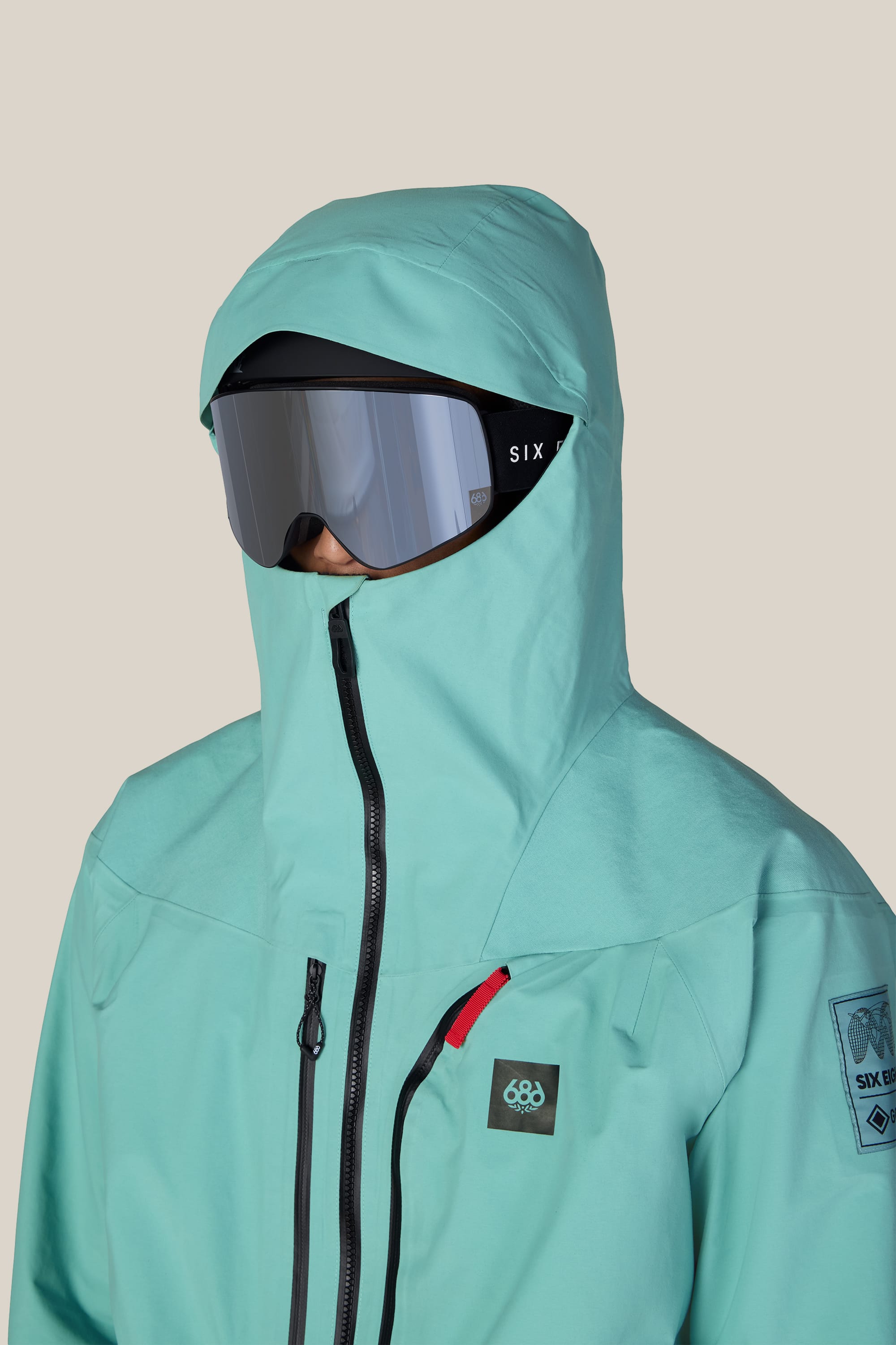 A person wearing the 686 Men’s GORE-TEX® Dispatch™ Thermagraph® Jacket in teal, hood up, with a black zipper and large reflective ski goggles. This 686 woven nylon jacket covers most of their face for added protection.