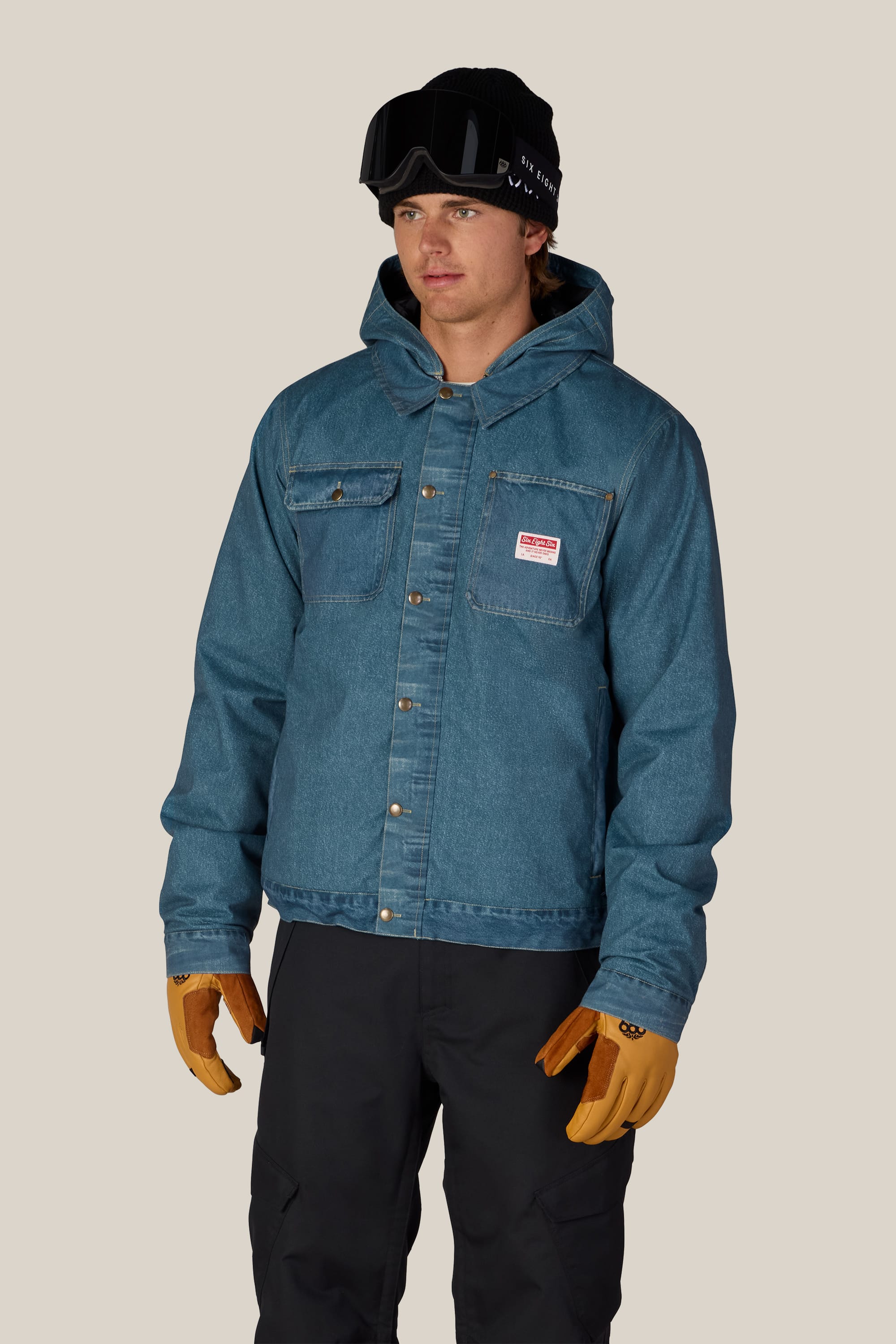 A person wearing a blue 686 Men’s Deconstructed™ Denim Insulated Jacket by 686, black snow goggles, tan gloves, and black pants stands against a plain beige background.