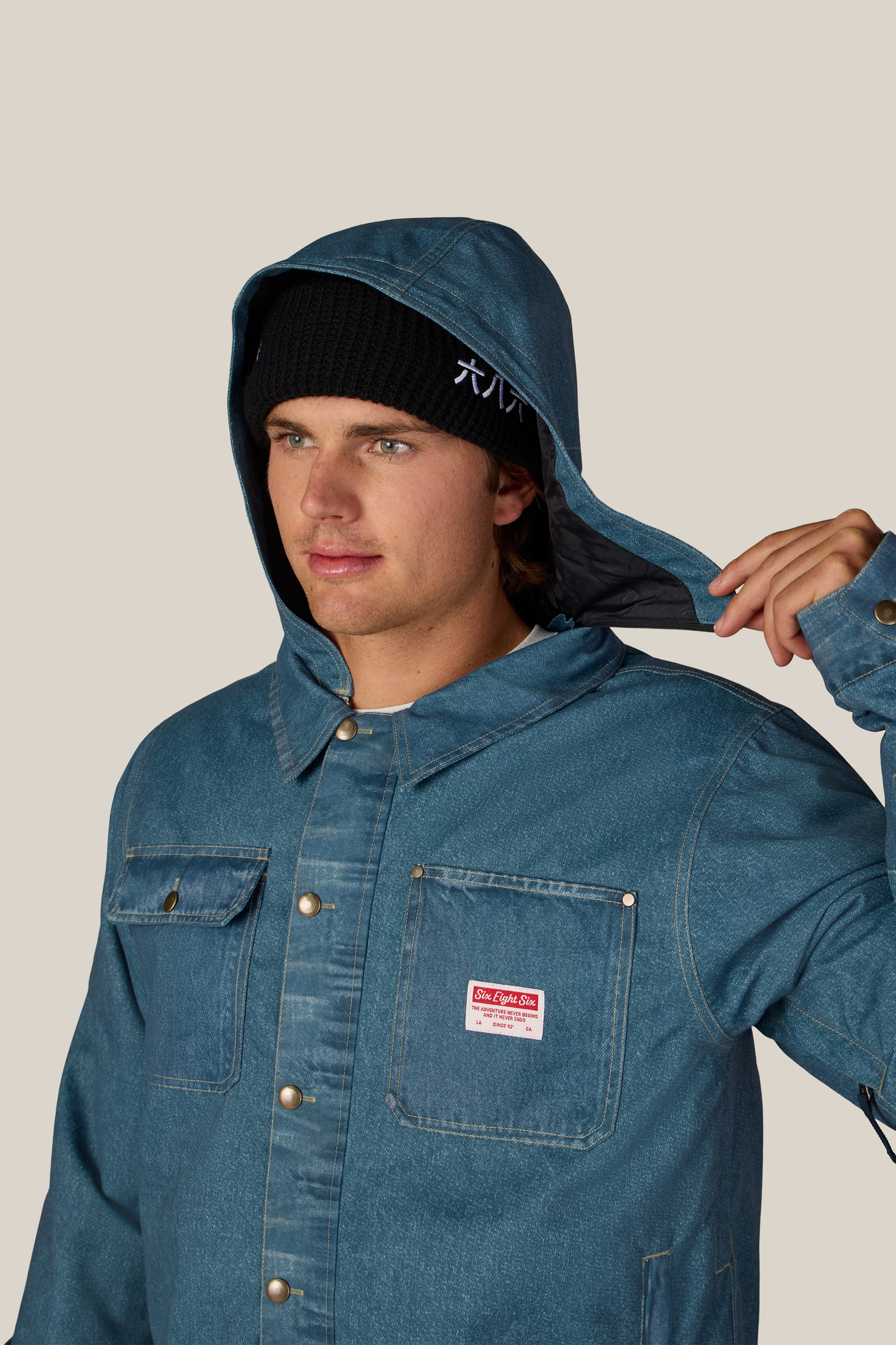 A person wearing the 686 Men’s Deconstructed™ Denim Insulated Jacket and a black beanie adjusts the jacket’s hood with one hand. The 686 denim jacket features metal buttons and a red and white label on the chest. The background is plain and light-colored.
