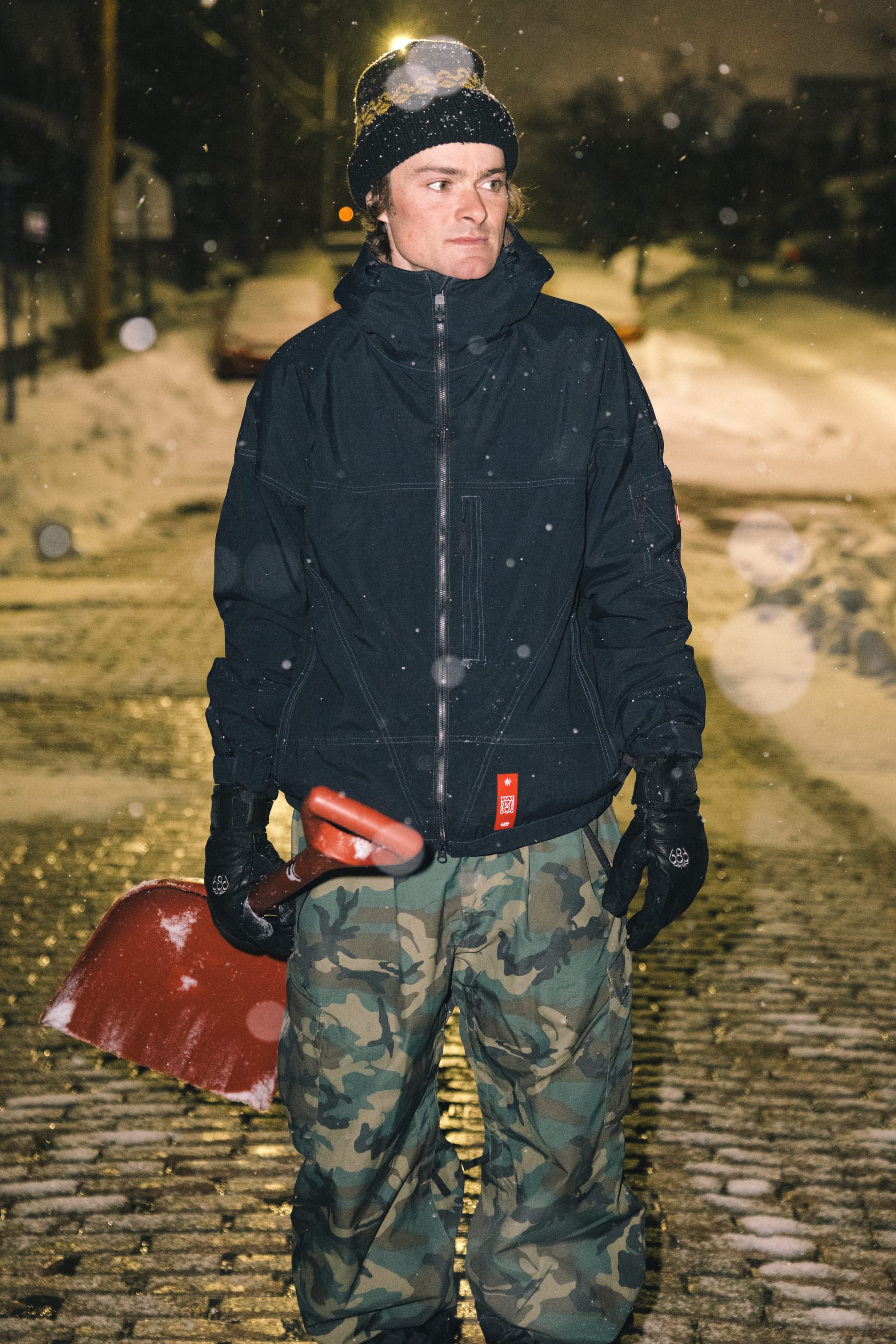 Wearing the 686 Men’s Dojo® Shell Jacket by 686 and camouflage pants, a person stands on a snowy street at night, holding a red snow shovel as snow falls around them.