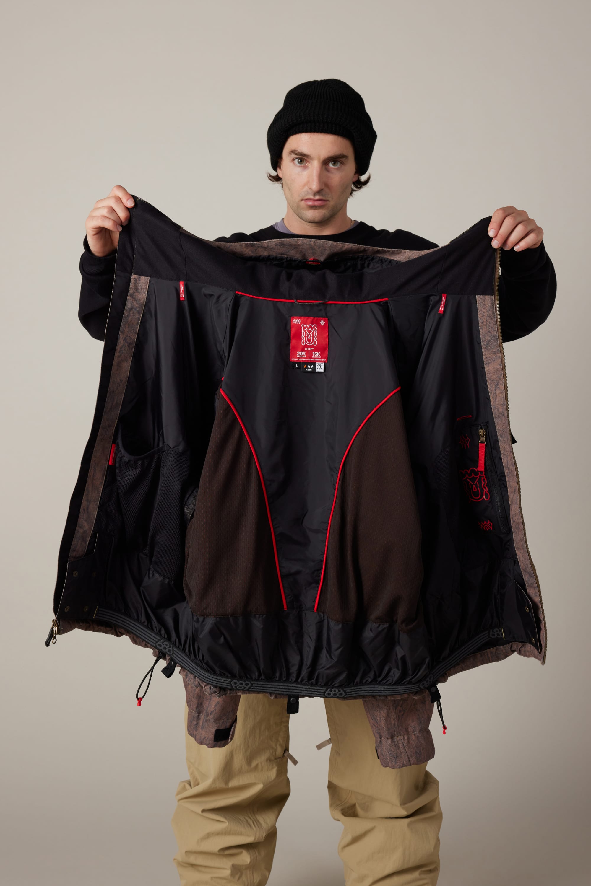 A man in a black beanie and beige pants holds open the 686 Men’s 686 Dojo® Shell Jacket by 686, showing its inner lining and several red tags, against a plain background.