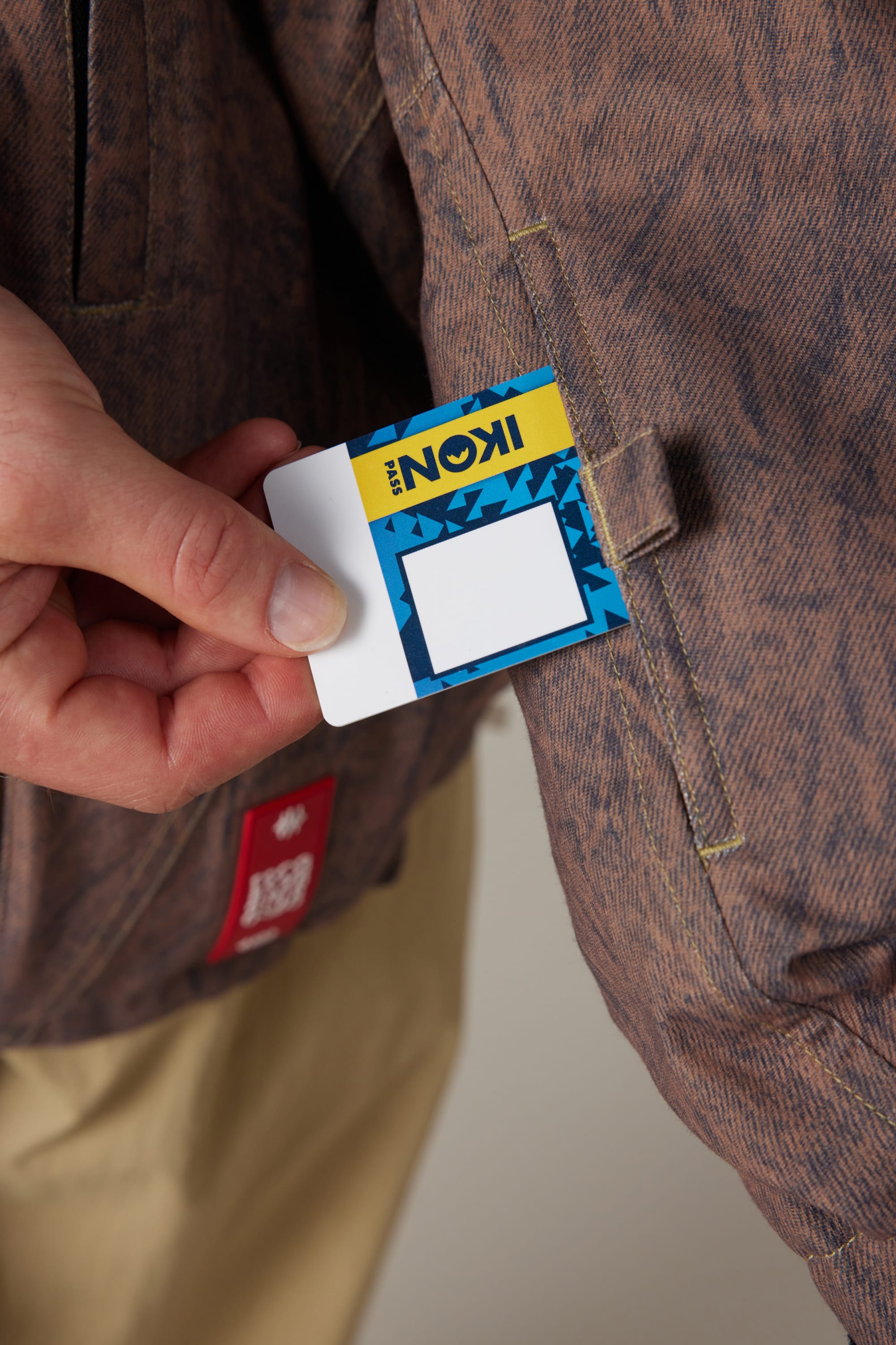 A hand pulls a blue and yellow IKON badge with a blank white square from the sleeve pocket of the 686 Men’s 686 Dojo® Shell Jacket. The brown waterproof jacket by 686 features a red tag for added detail.
